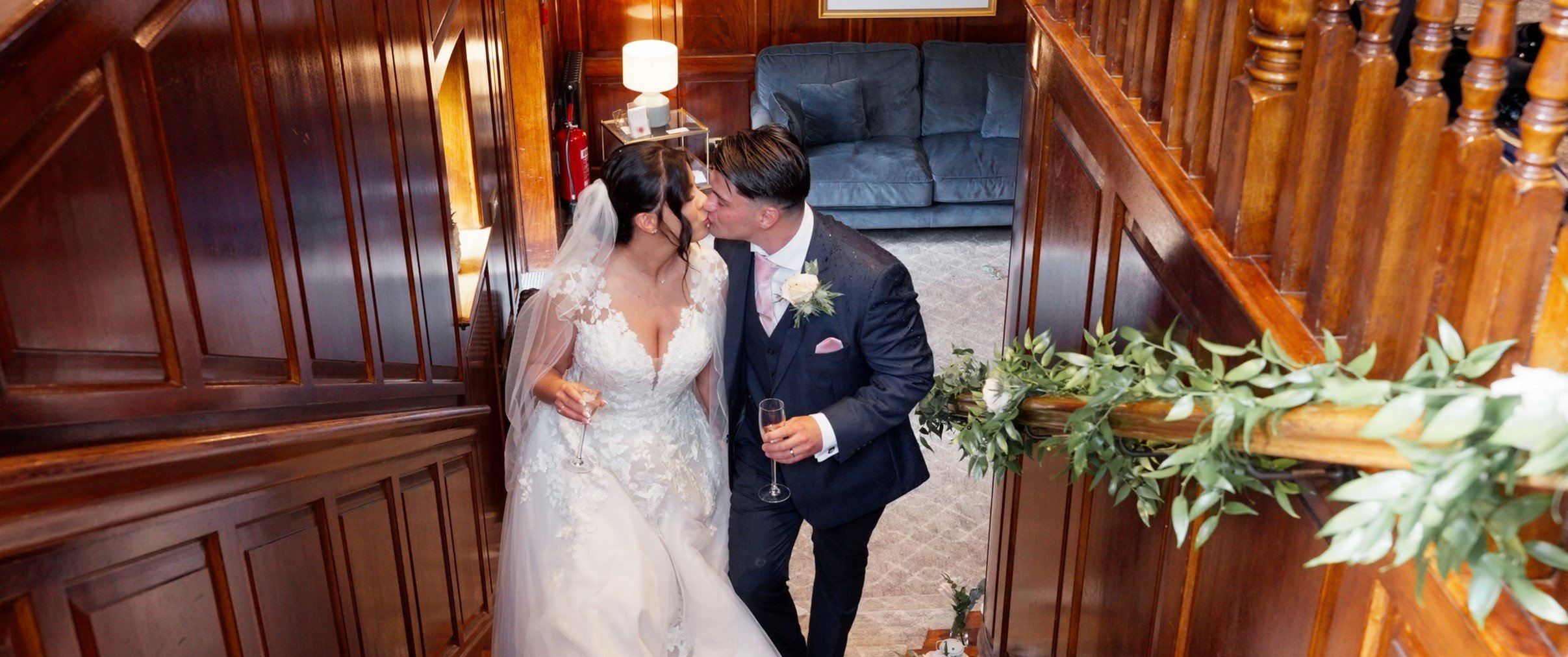 A bride and groom sharing a kiss at their wedding, holding glasses of champagne, in a wood-paneled staircase area with a blue couch and a small side table in the background.