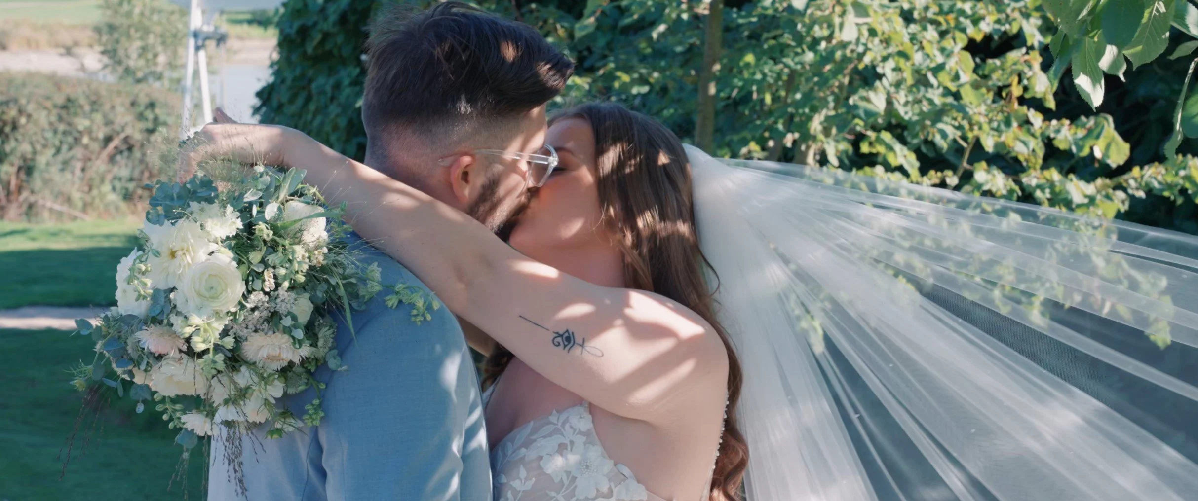 A bride and groom share a kiss outdoors during their wedding, with the groom carrying a bouquet of white and green flowers. The bride's long veil is flowing behind her, and sunlight filters through the trees in the background.