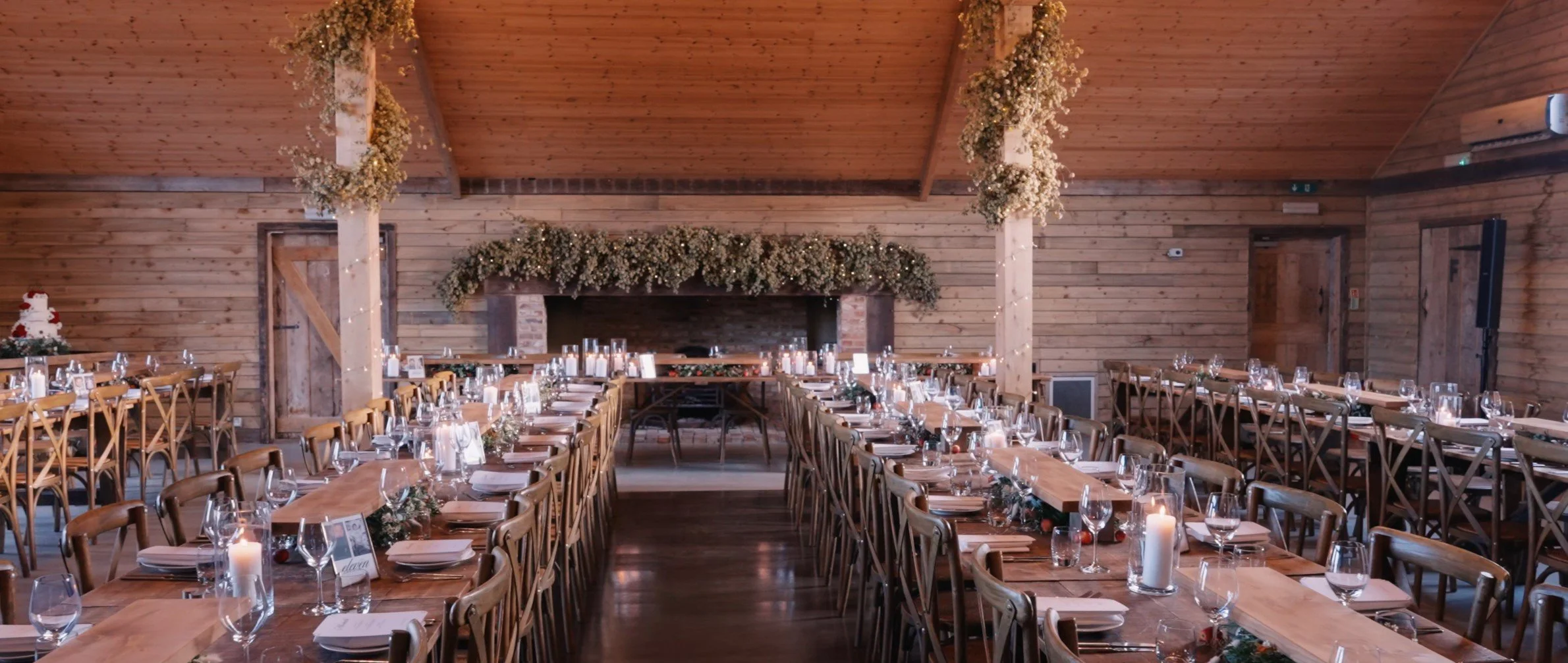 Wedding reception decorated with wooden tables and chairs, white candles, and floral arrangements in a rustic barn with wood-paneled walls and ceiling.