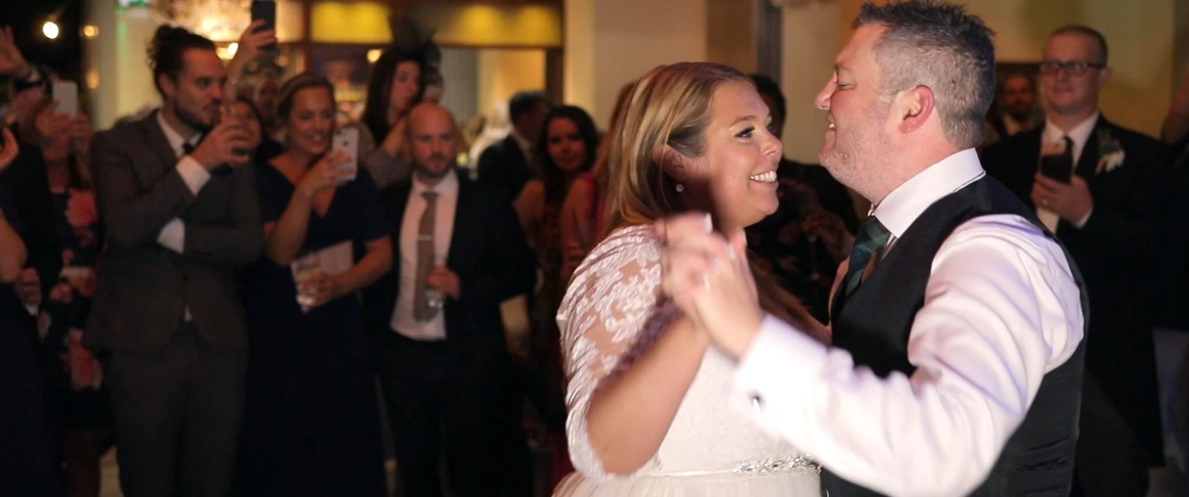 A bride and groom dancing closely together at their wedding reception while guests watch and take photos.