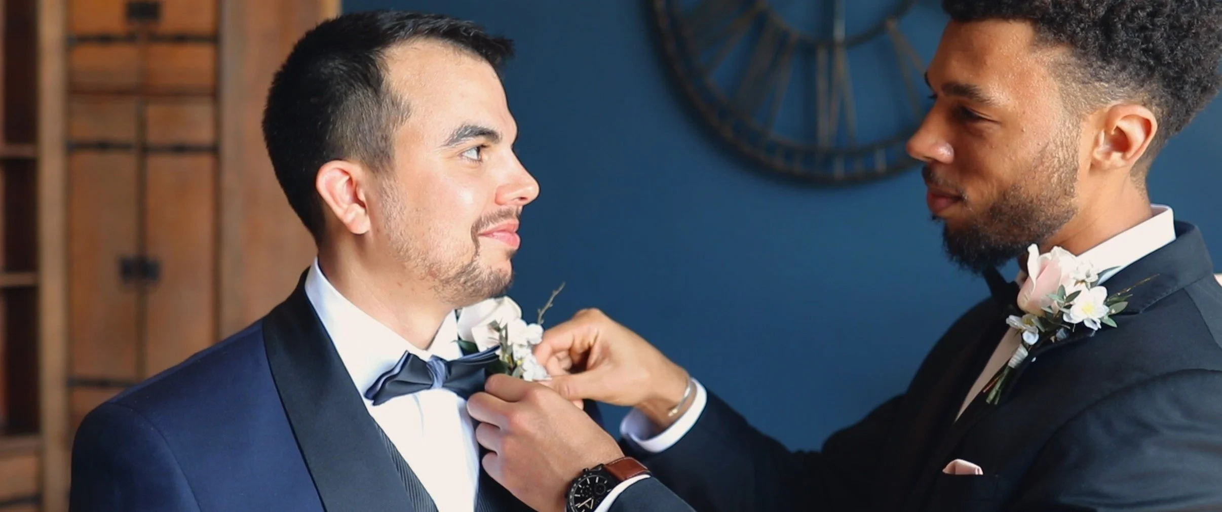 Two men in tuxedos, one helping the other adjust his bow tie before a wedding ceremony, with floral boutonnieres on their lapels.