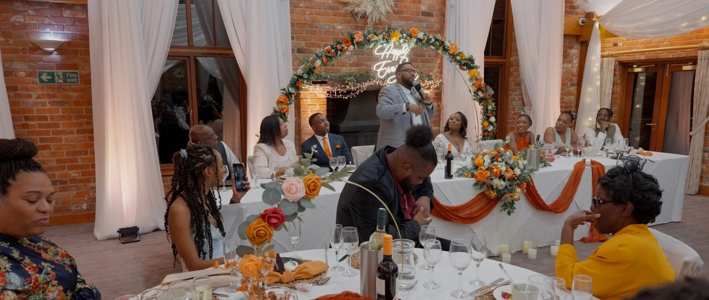 A speaker giving a toast or speech at a wedding reception, with guests seated at tables decorated with flowers and candles, and a floral arch with a "Happy End" sign behind him.