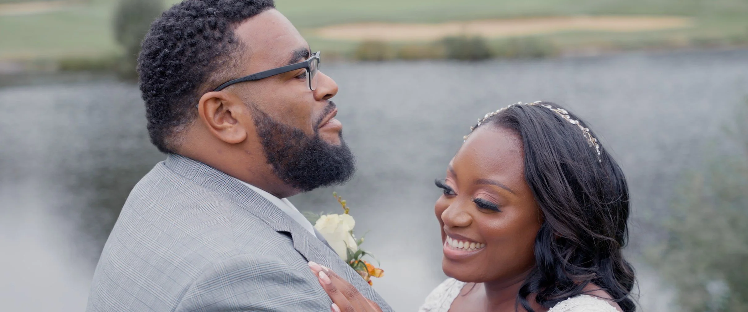A couple on their wedding day, smiling and embracing outdoors by a body of water.