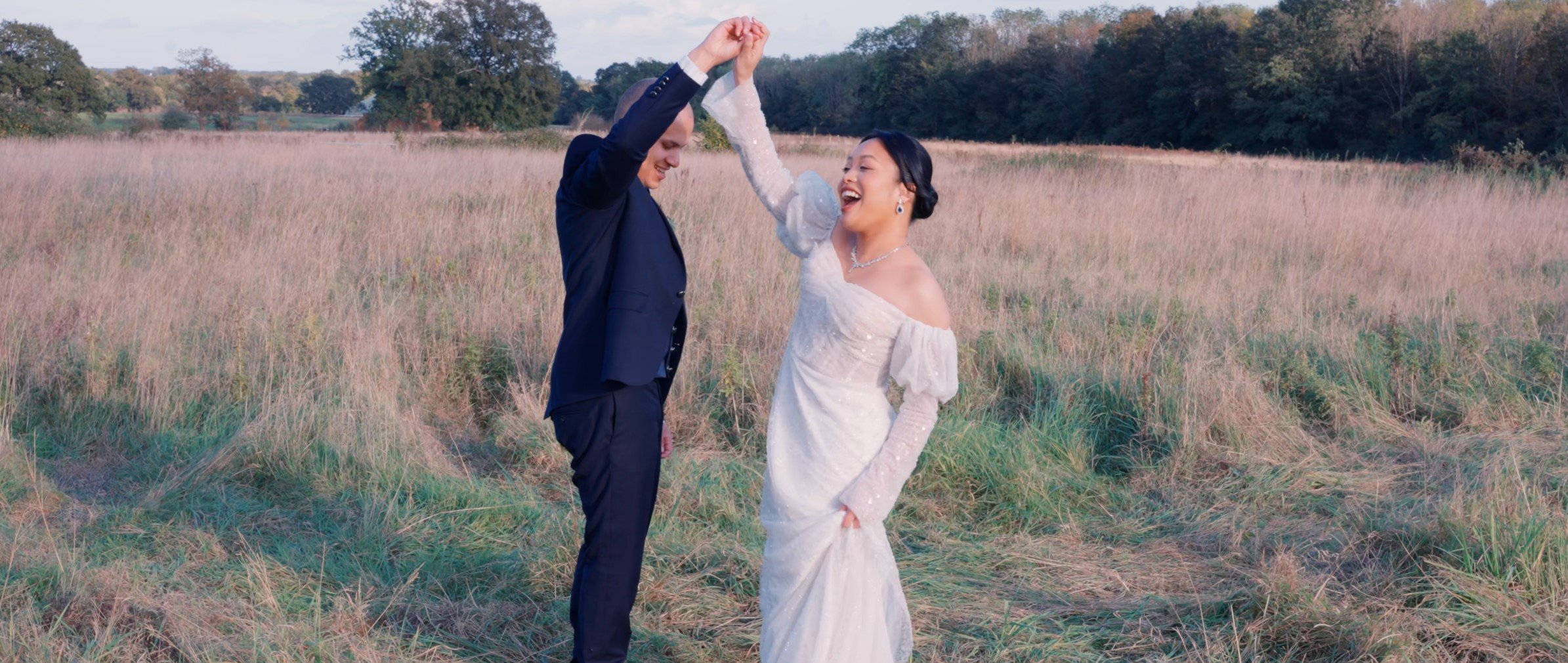 A happy couple in wedding attire dancing in an open grassy field with trees in the background during late afternoon or early evening.