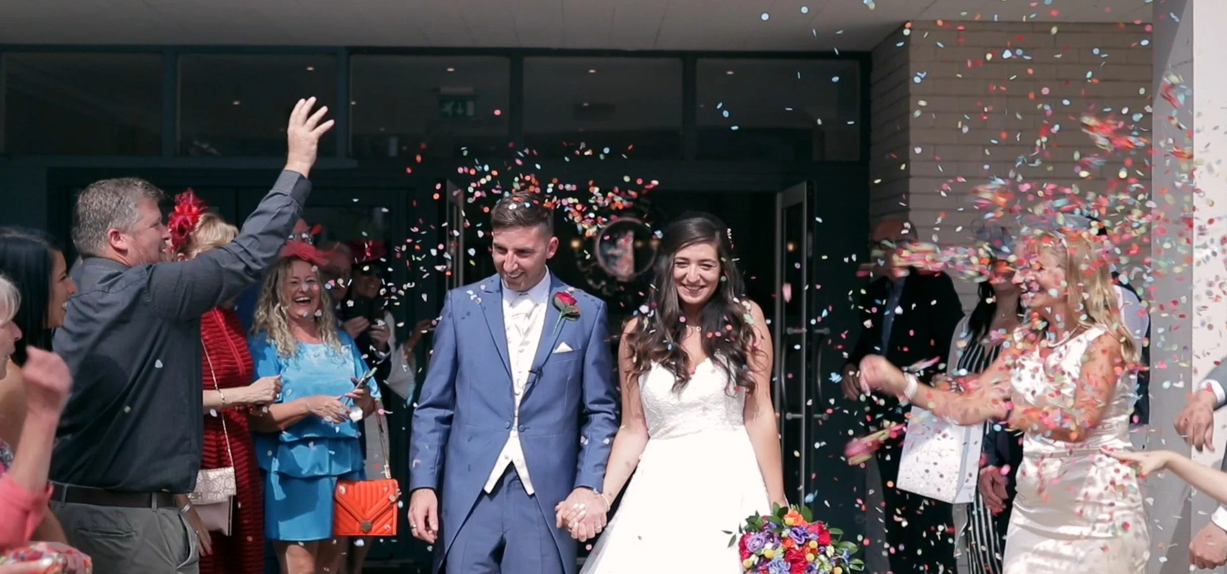 A newlywed couple holding hands exiting a building as confetti falls around them, surrounded by friends and family celebrating.