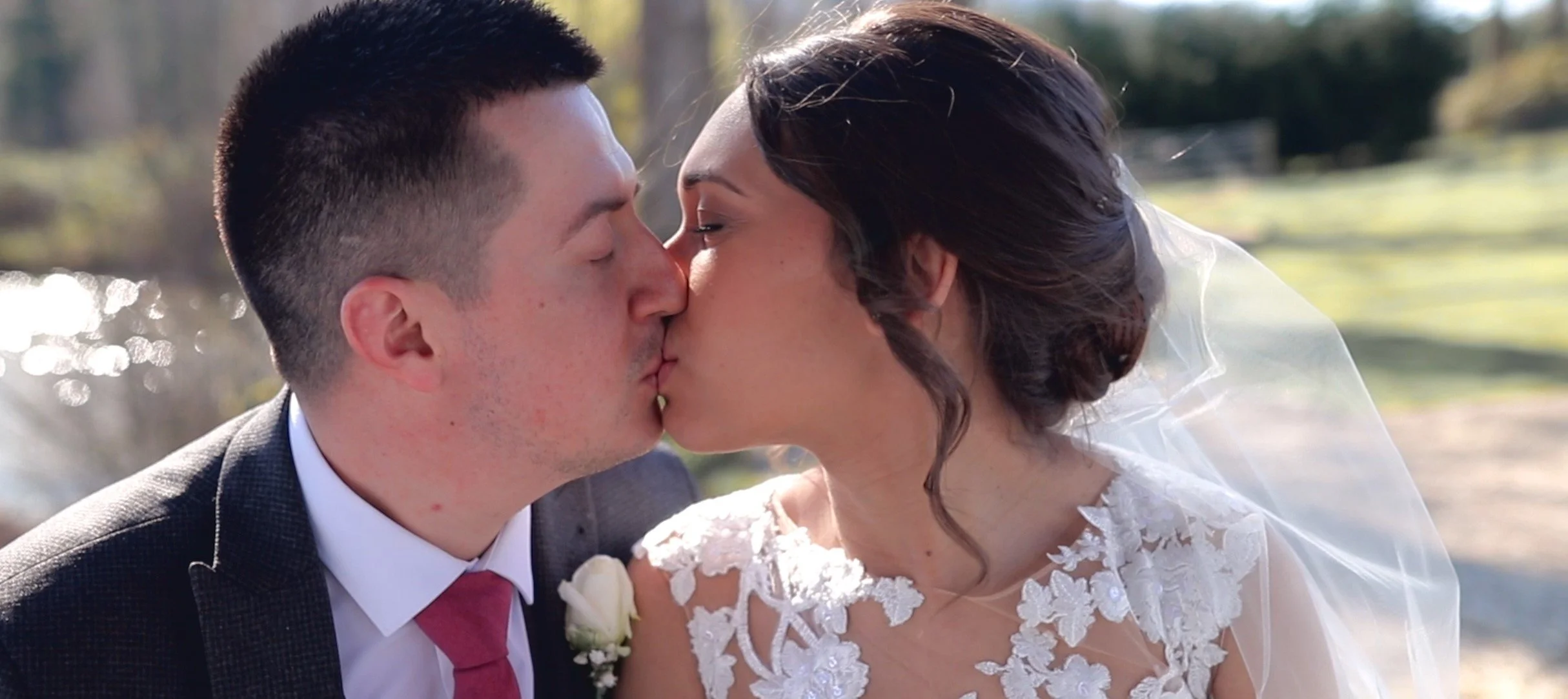 A bride and groom sharing a kiss outdoors during their wedding, with sunlight and greenery in the background.