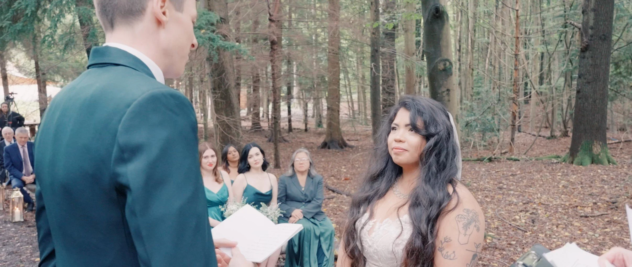 A wedding ceremony taking place outdoors in a wooded area with a bride and groom exchanging vows, surrounded by seated guests and a forest background.