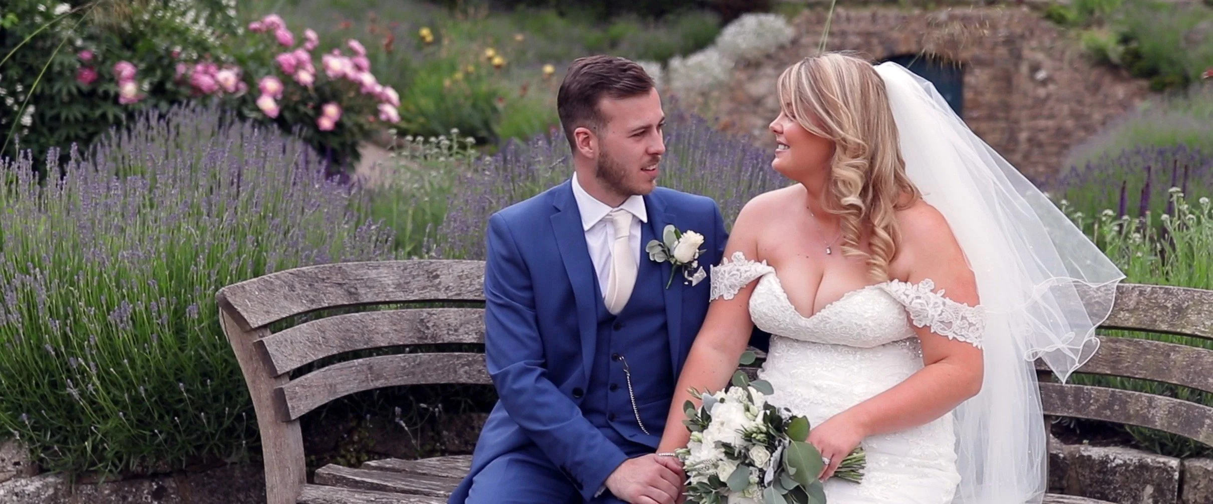 A bride and groom sitting on a wooden bench in a garden with purple and pink flowers, smiling and looking at each other. The bride is in a white lace wedding dress with a veil, holding a bouquet of white flowers and greenery. The groom is in a blue s