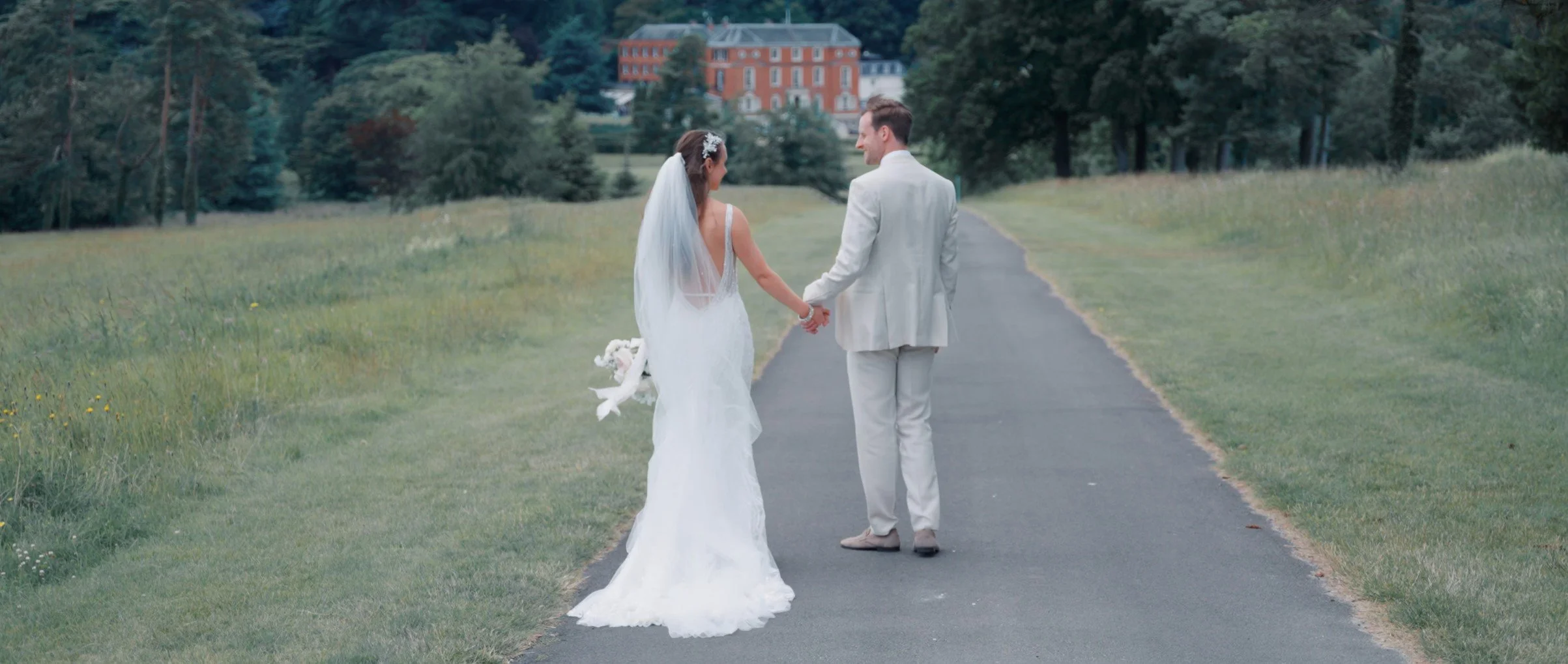 A bride and groom walking hand in hand on a paved path through a grassy park-like area with trees and a large building in the background.