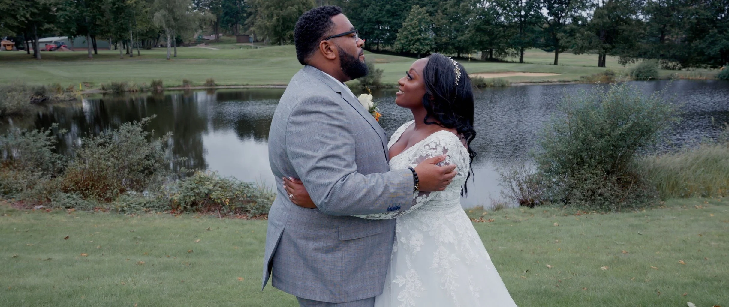 A newlywed couple standing close together outdoors near a pond, embracing and looking lovingly at each other. The groom is wearing a light gray suit and glasses, and the bride is dressed in a white lace wedding gown with dark wavy hair and a headband