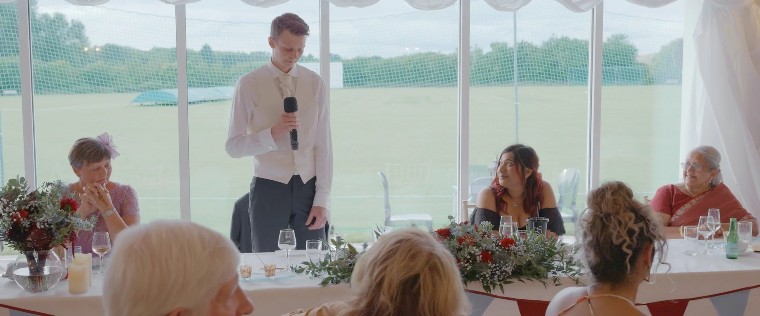 A young man giving a speech at a wedding reception, standing behind a decorated table with floral arrangements, while guests sit and listen inside a large tent with panoramic windows and a view of a green field and trees outside.