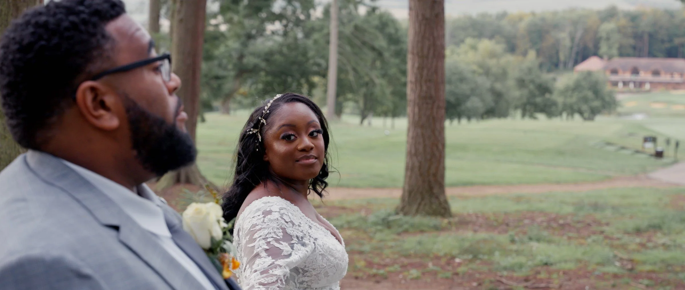 Bride in a white lace wedding dress looking at camera, groom in a light gray suit and glasses standing nearby, in an outdoor park setting with trees and a large building in the background.