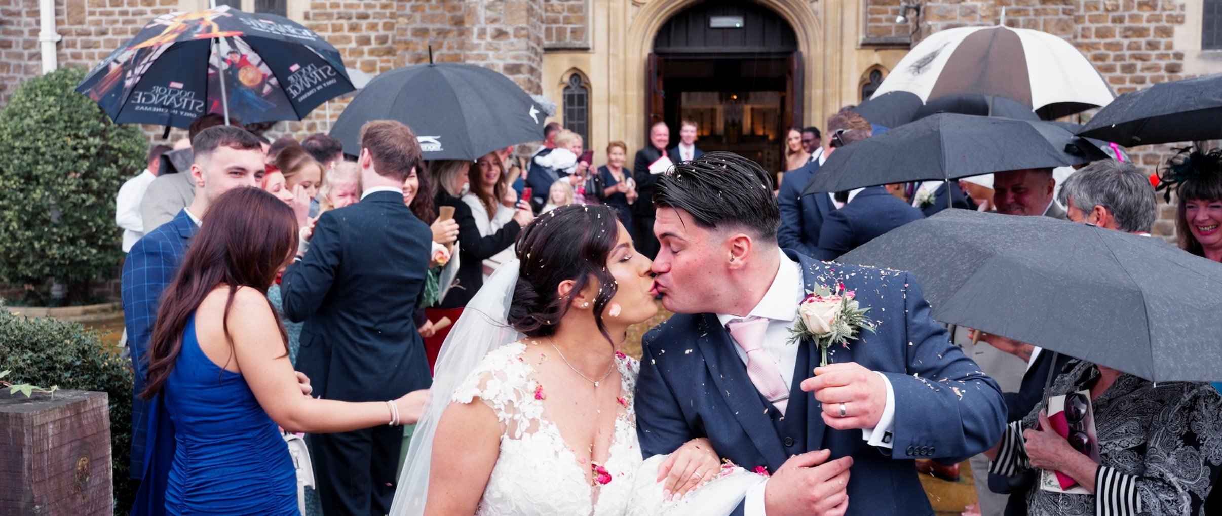 Bride and groom kissing under umbrellas outside a church, surrounded by wedding guests at a rainy wedding celebration.