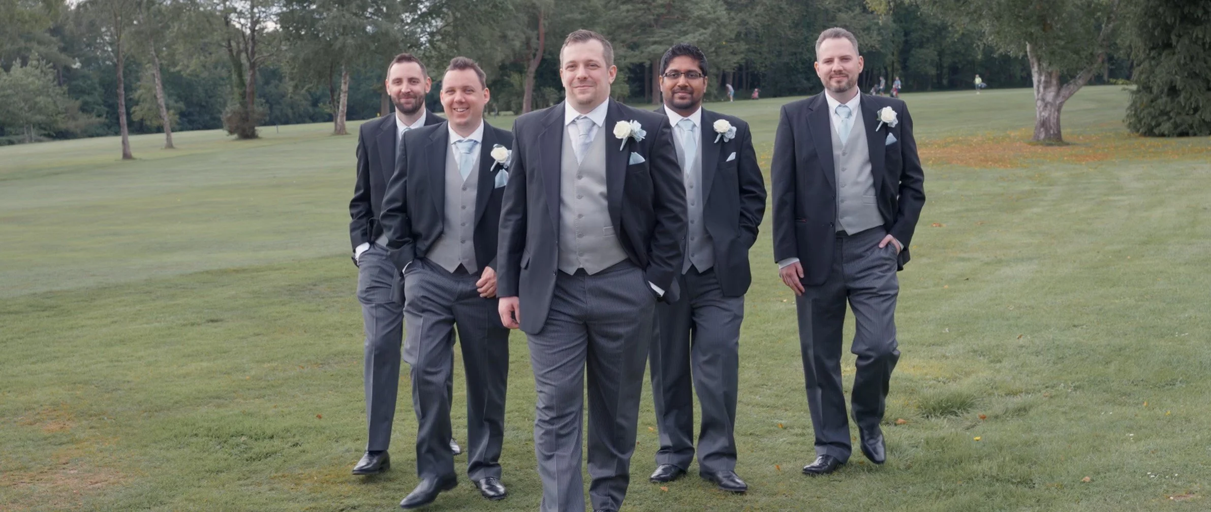 Group of six men dressed in tuxedos and suits walking together on a green grassy field, with trees in the background, at a wedding or formal event.