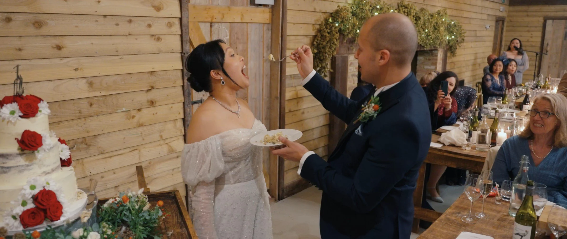 A bride with dark hair in an off-shoulder white wedding dress is being fed cake by the groom, who is wearing a dark suit with a boutonniere, at a wedding reception with wooden walls and guests seated at tables.