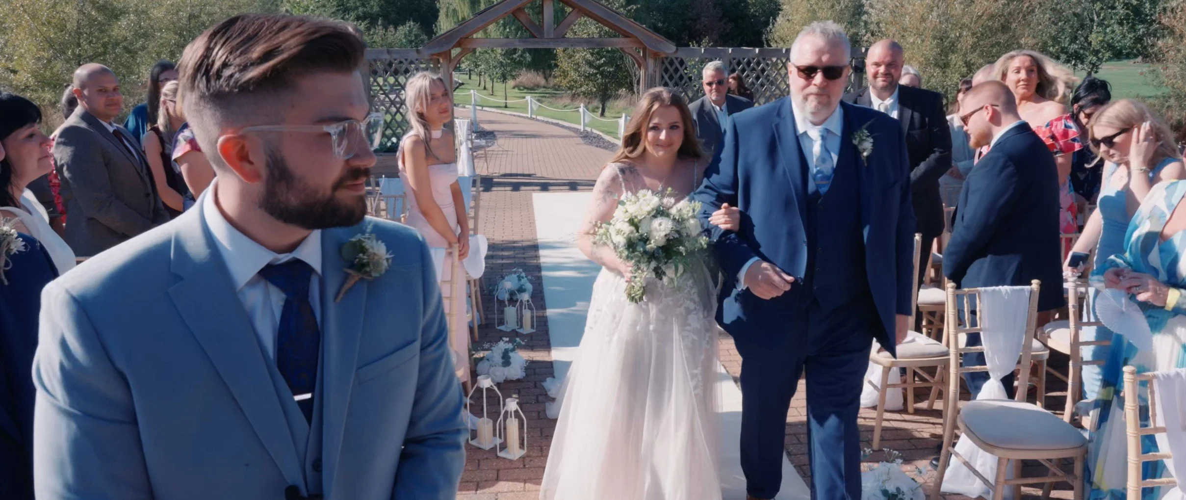 A wedding ceremony outdoors with the bride, walking arm-in-arm with an older man, likely her father, down the aisle lined with chairs and floral decorations. Guests are standing and seated, watching the procession, on a sunny day in a garden setting.
