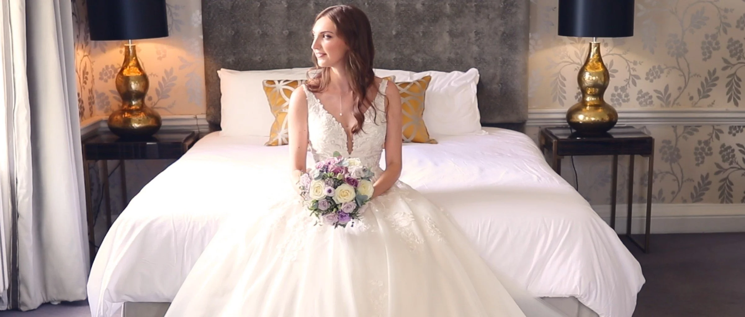A woman in a wedding dress sitting on a bed holding a bouquet of flowers in a bedroom with two gold lamps on bedside tables.