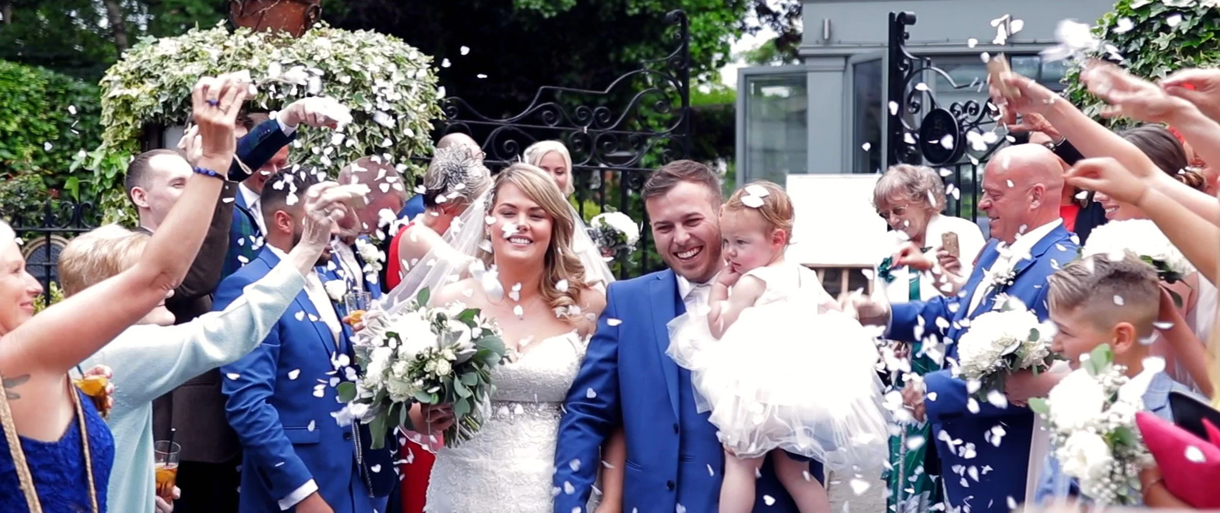 A newlywed couple in wedding attire walking through a crowd of family and friends throwing white flower petals, celebrating outdoors.
