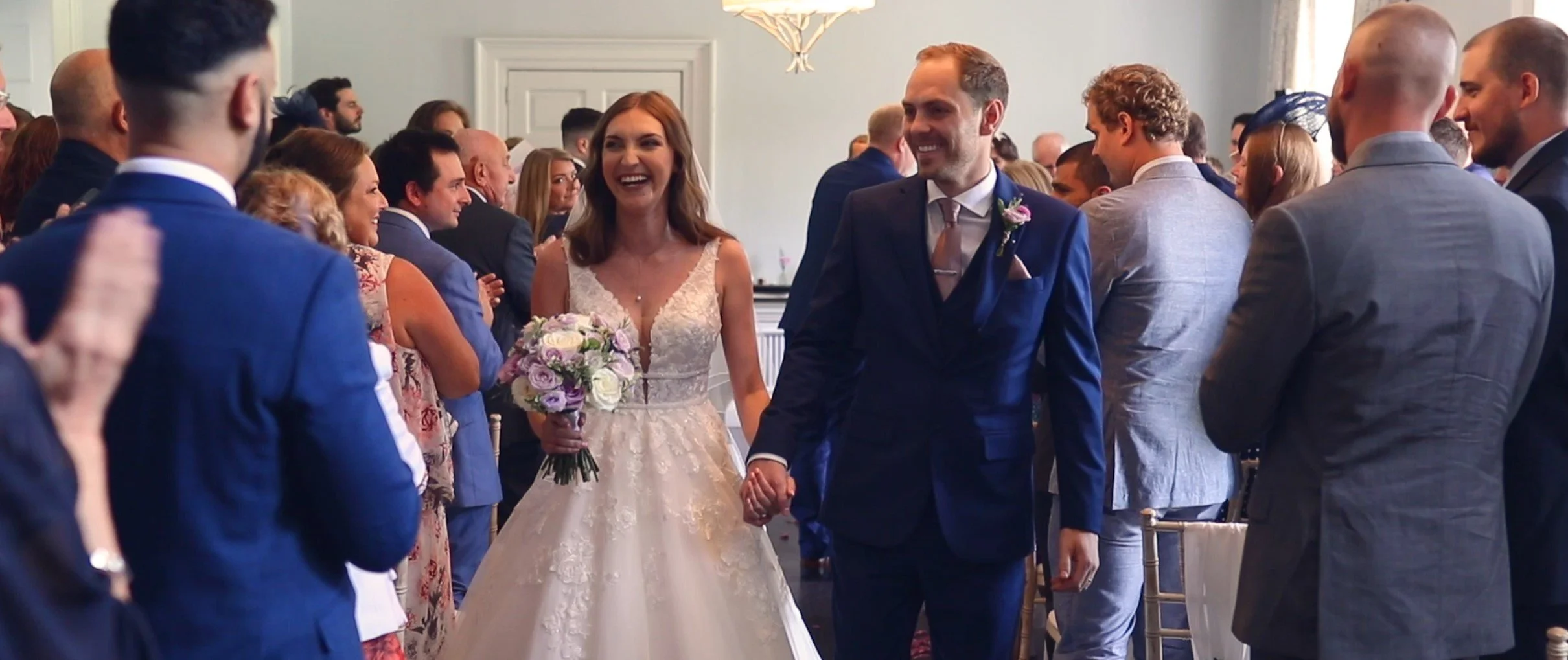 A bride and groom walking hand in hand through a room filled with wedding guests who are smiling and clapping, celebrating their wedding.
