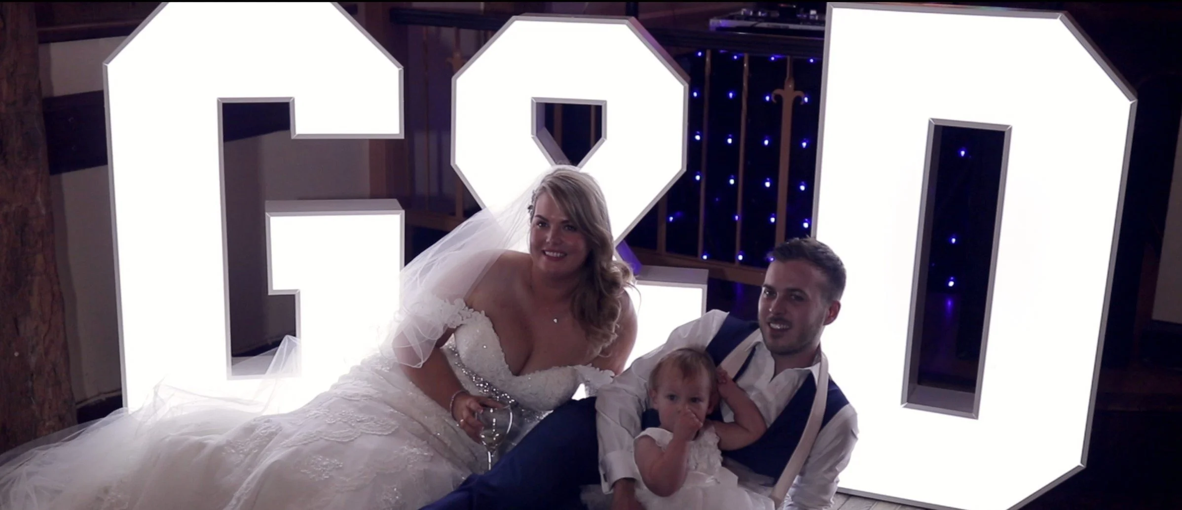 A family of three at a wedding, with large illuminated letters spelling 'LOVE' behind them, including a bride in a wedding dress, a groom in a vest and shirt, and a young girl in a white dress.