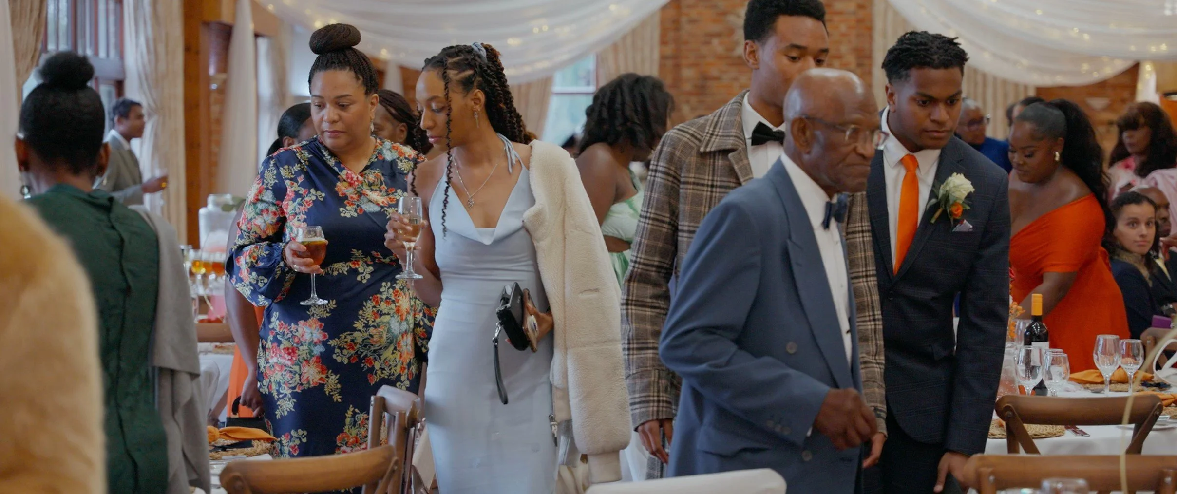 People standing in line at a banquet or wedding reception, dressed in formal and semi-formal attire, holding drinks, in a decorated indoor venue.