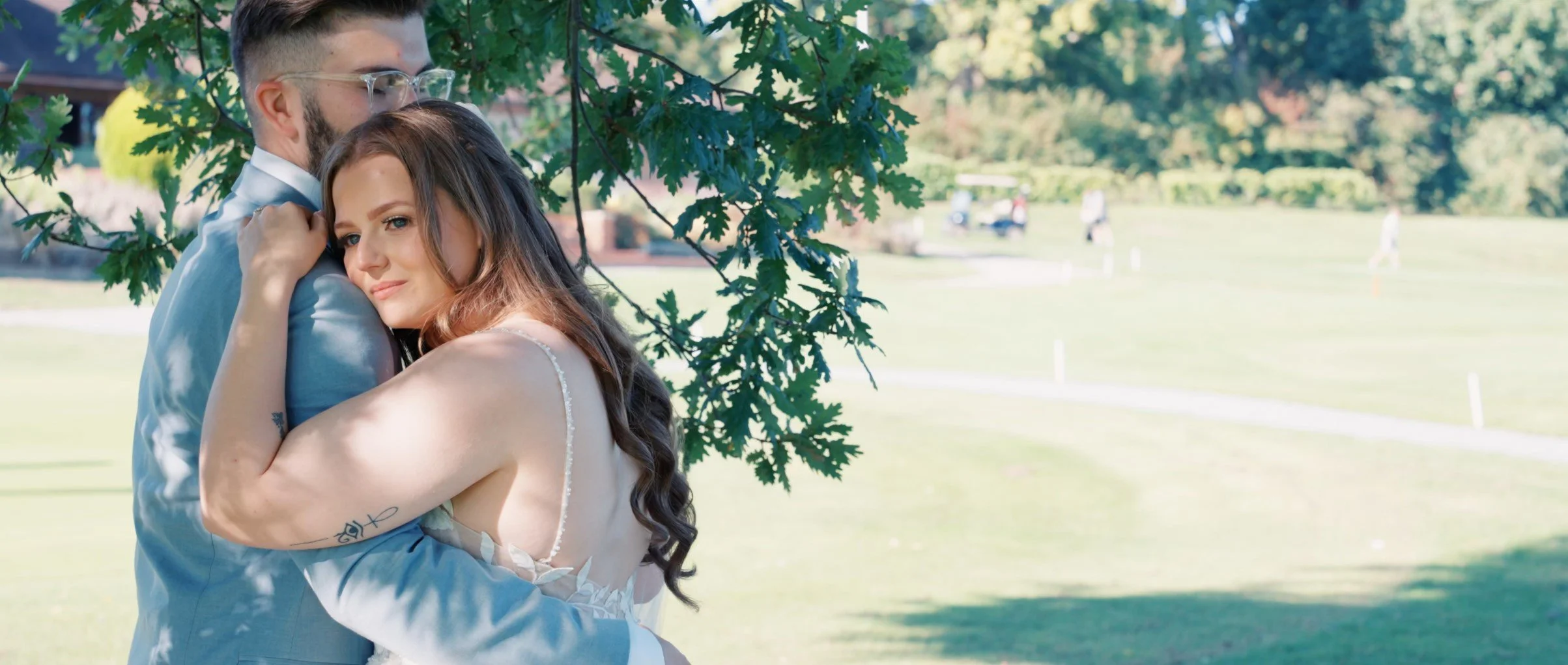 A couple embraces under a tree in a park, with the woman resting her head on the man's shoulder and both showing content expressions, surrounded by greenery and a sunny outdoor setting.