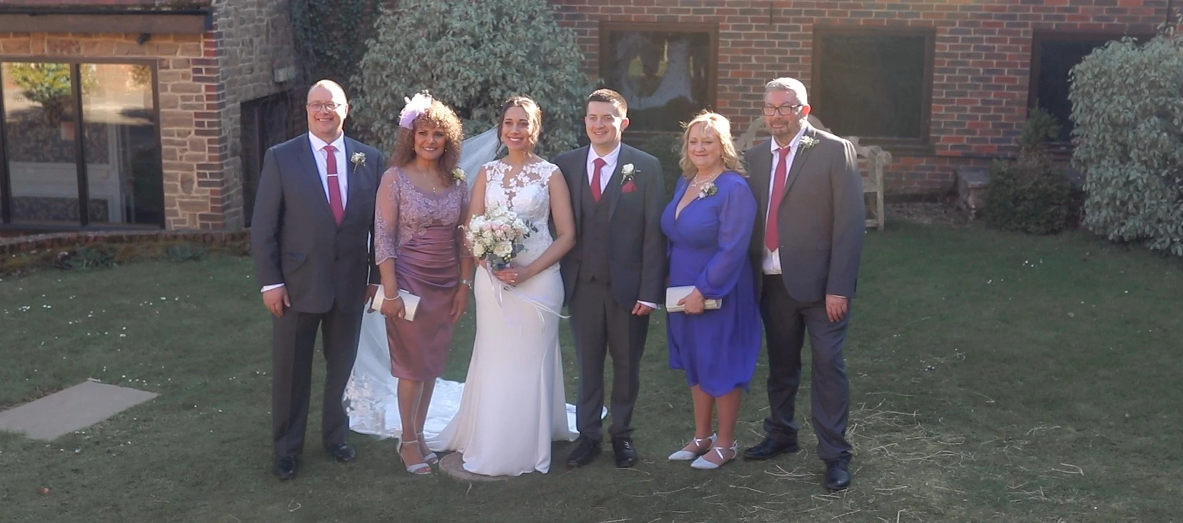 Group of seven people standing outdoors in front of a brick building, dressed in formal attire for a wedding.