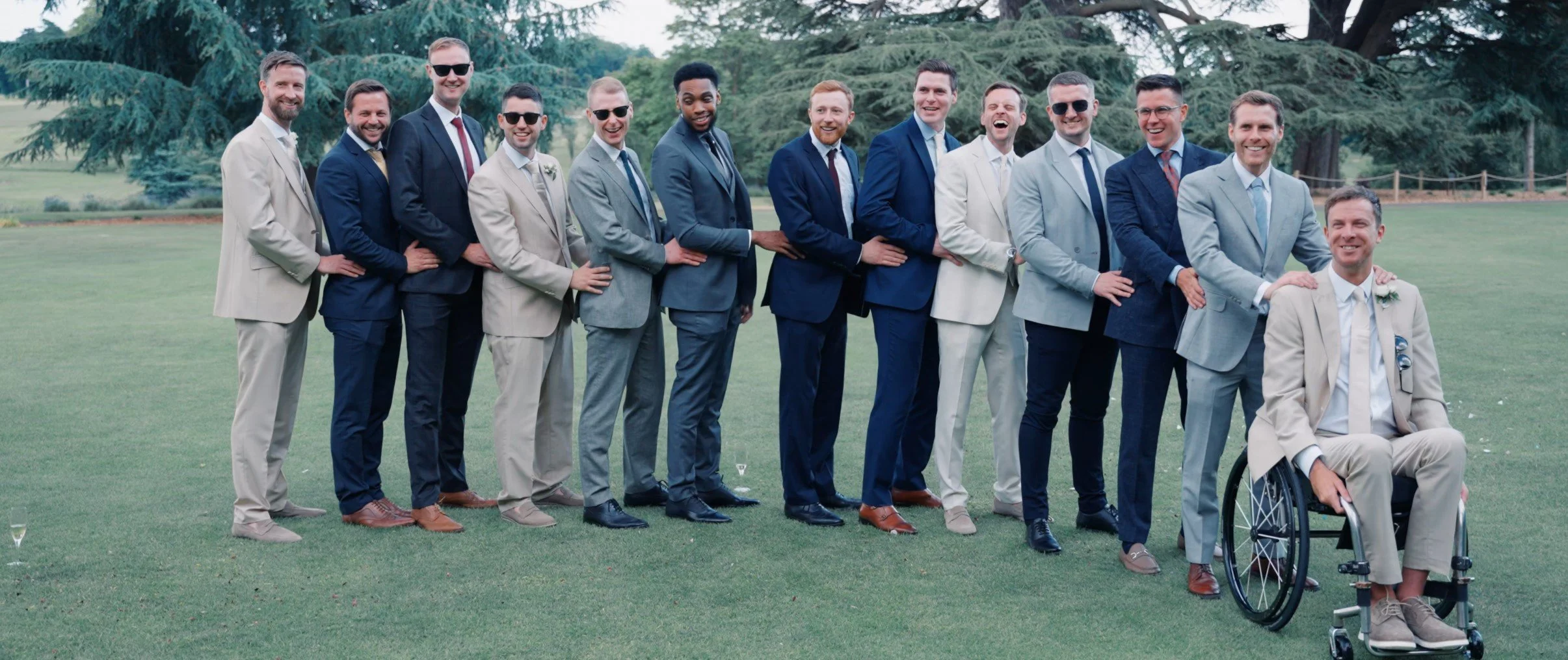 A group of men dressed in suits standing in a line outdoors on a grassy area with trees in the background, one man seated in a wheelchair.