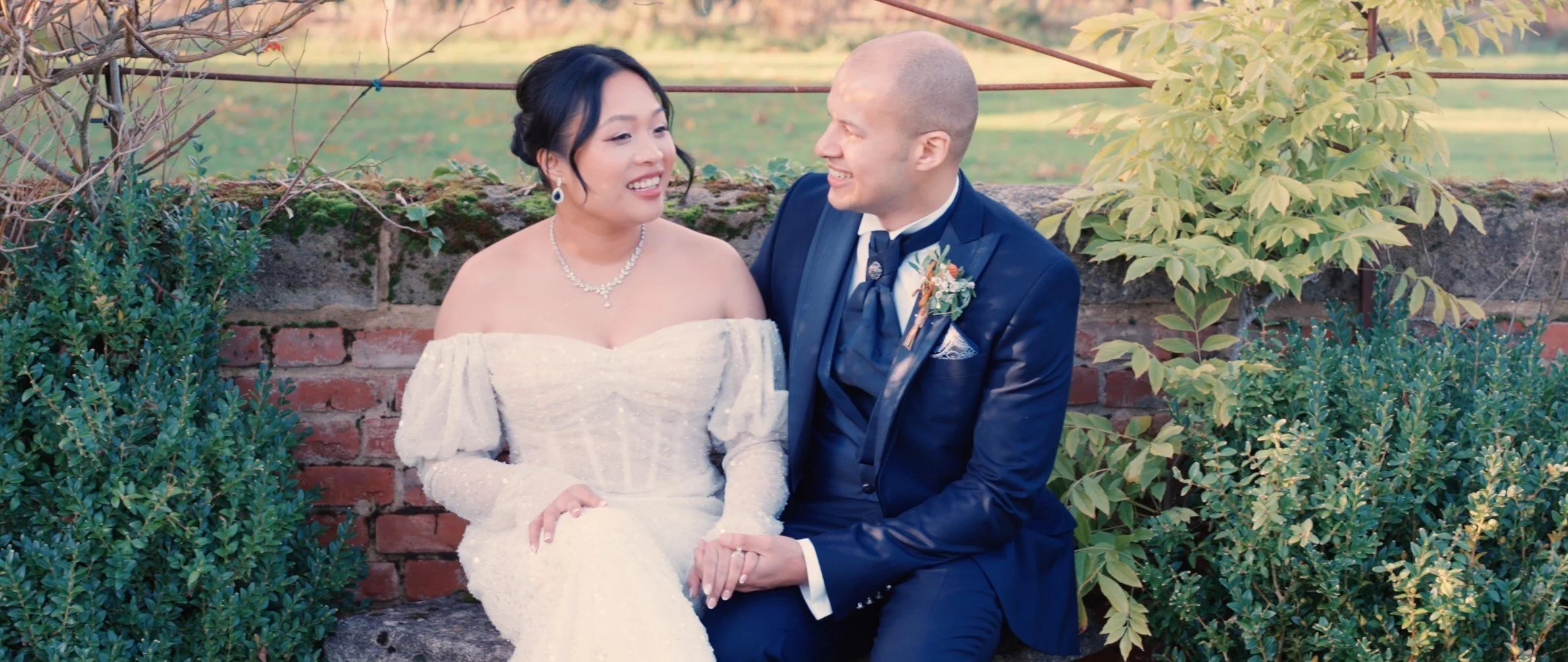 A newlywed couple seated on a stone bench outdoors, smiling and holding hands. The bride is wearing a white off-the-shoulder wedding dress with jewelry, and the groom is in a dark blue suit with a boutonniere. There are green bushes and a brick wall 