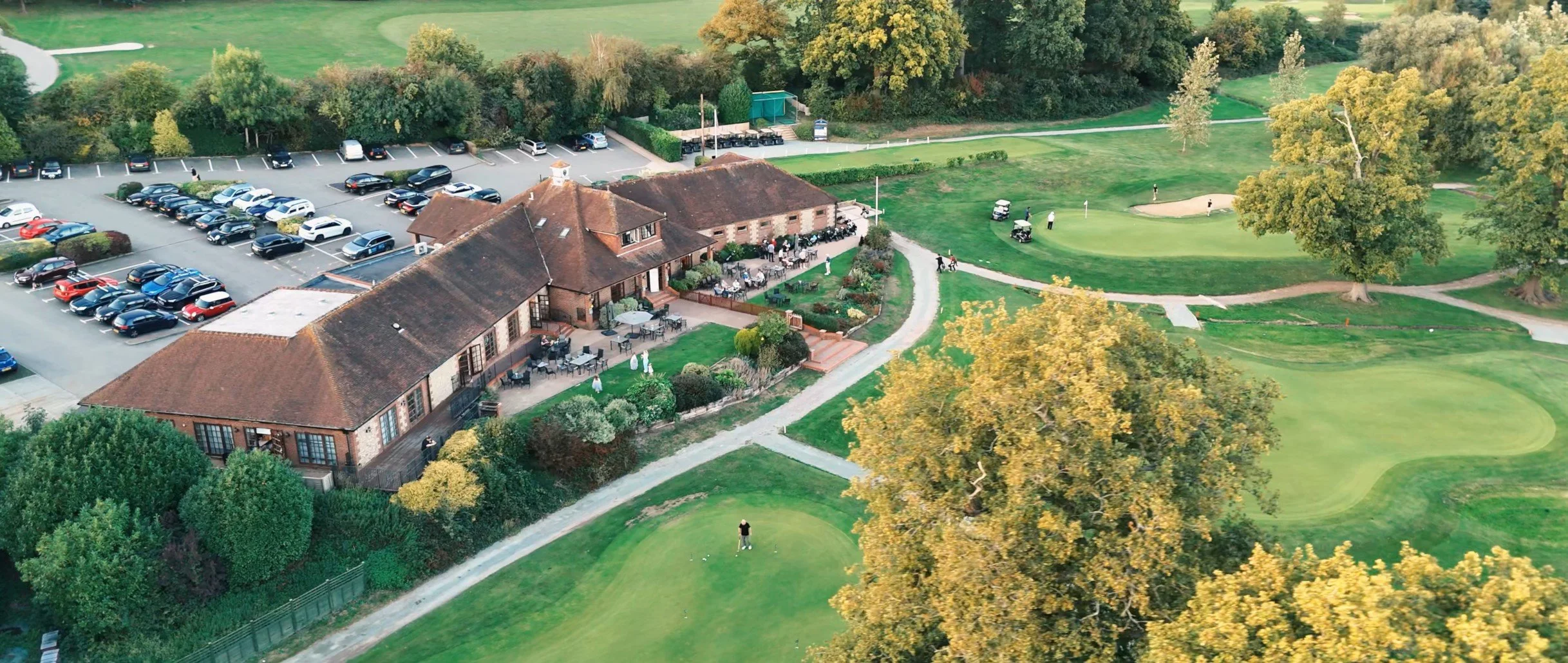 Aerial view of a golf course with several players, a clubhouse with outdoor seating, a parking lot filled with cars, and lush green trees surrounding the area.