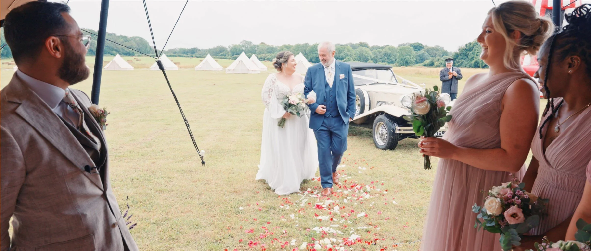 A wedding scene outdoors with a bride and a man walking down the aisle, surrounded by bridesmaids holding bouquets. There are tents in the background, and the ground is decorated with flower petals.
