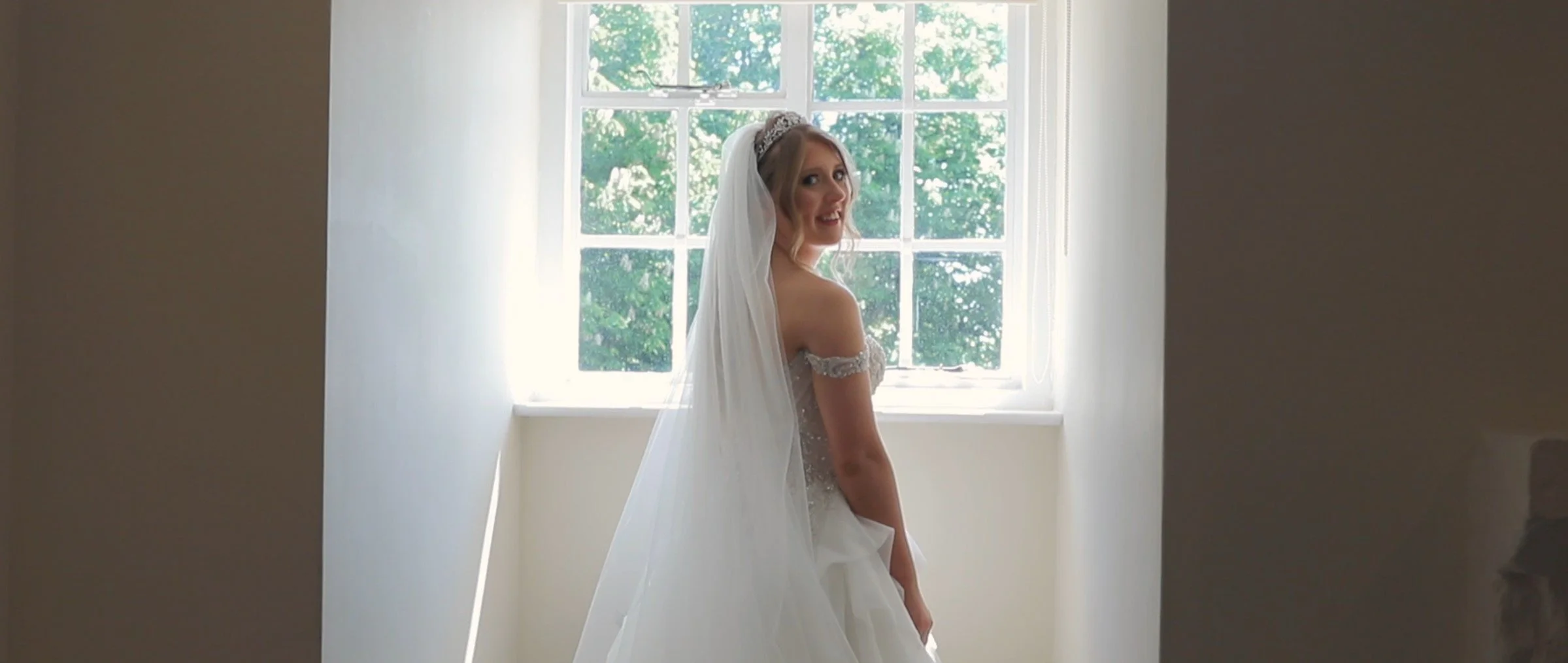 A bride in a wedding dress and veil standing in front of a window with greenery outside, looking over her shoulder and smiling.