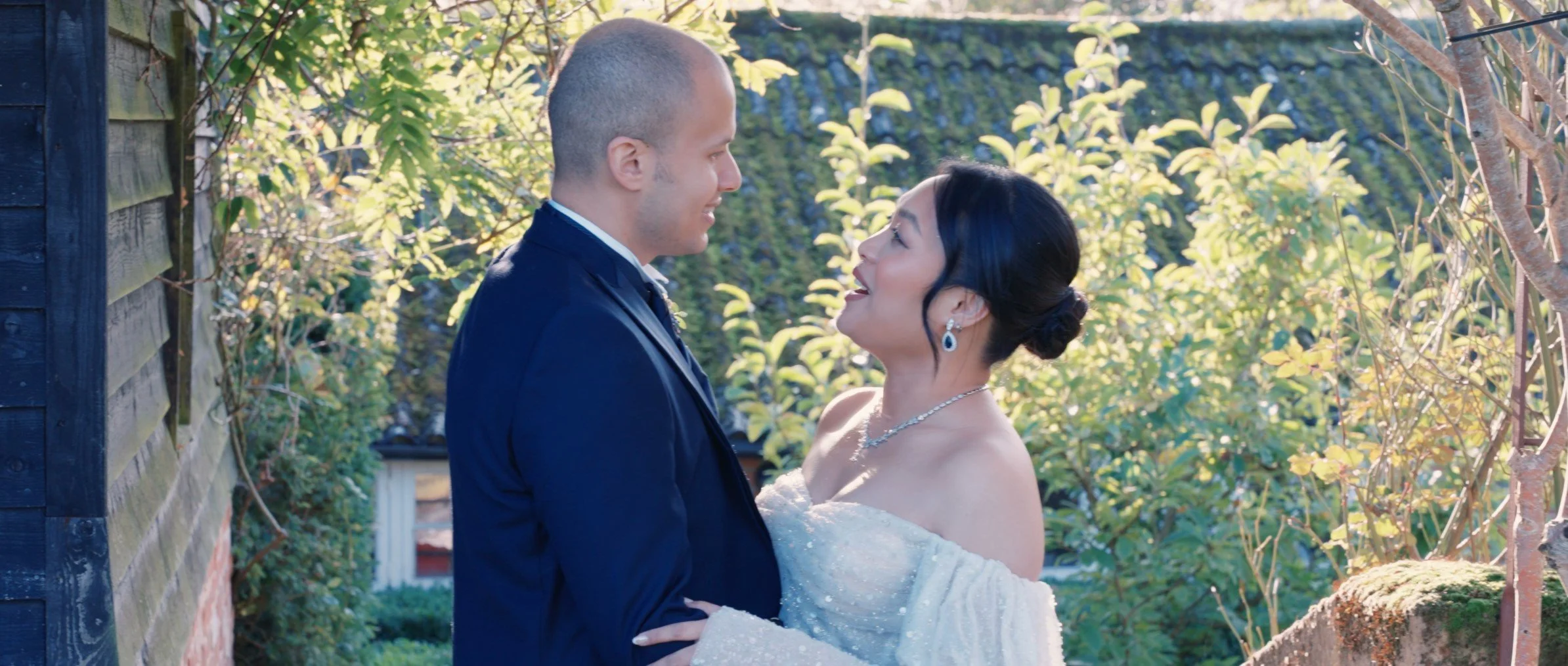 A newlywed couple sharing a joyful moment outside, with the groom in a dark suit and the bride in a white dress and jewelry, surrounded by greenery and sunlight.