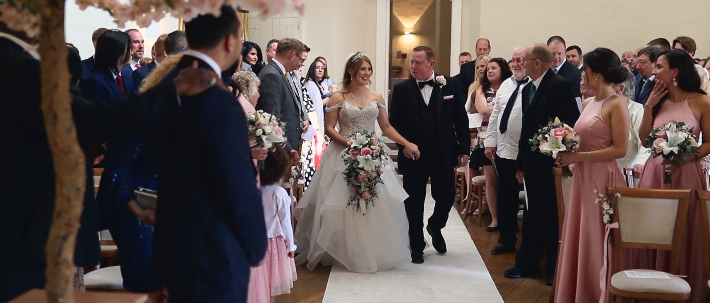 A bride in a wedding dress holding hands with a groom in a tuxedo, walking down the aisle at a wedding ceremony, surrounded by seated guests and bridesmaids in pink dresses holding floral bouquets.