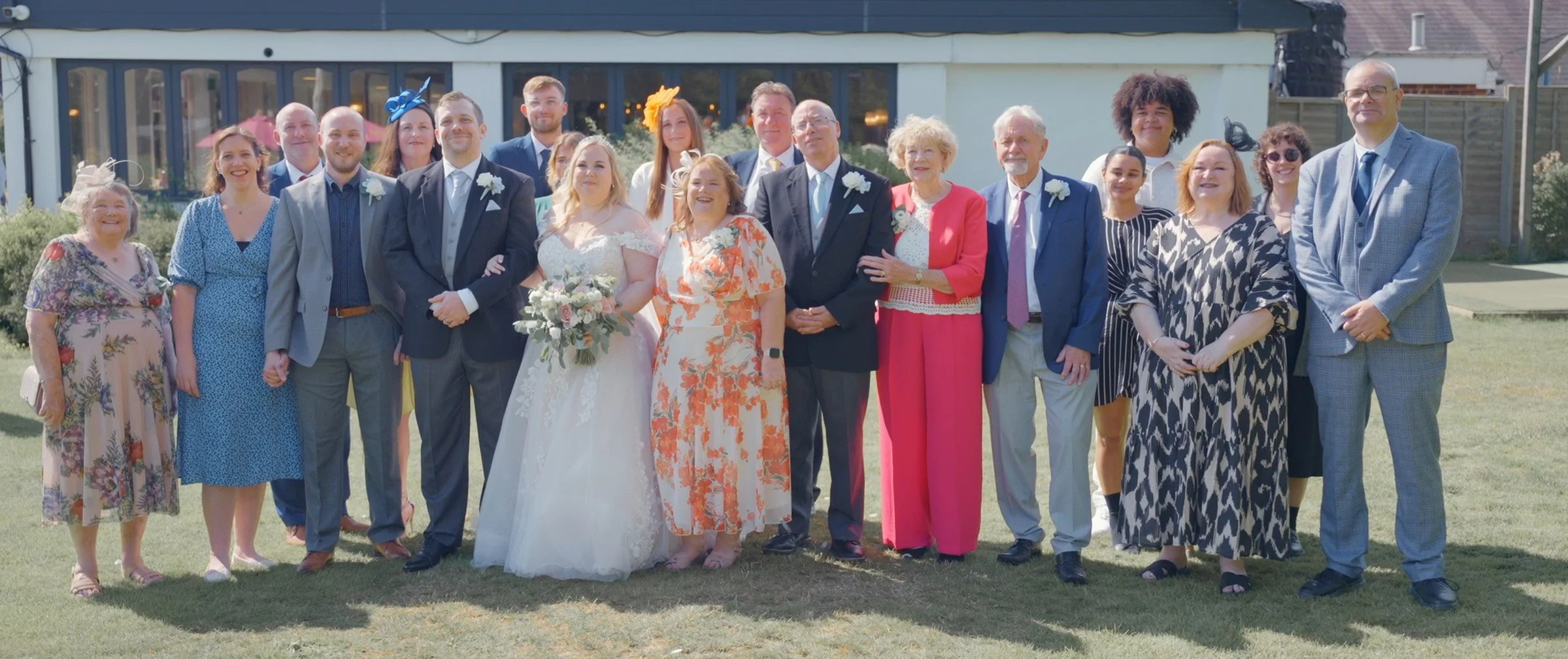 Group of people at wedding, standing outside on grass, with bride in white dress holding bouquet in center, groom in suit beside her, guests in colorful attire behind with buildings in background.