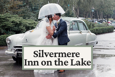 A bride and groom sharing a kiss under an umbrella in front of a vintage car, with a sign reading "Silvermere Inn on the Lake."
