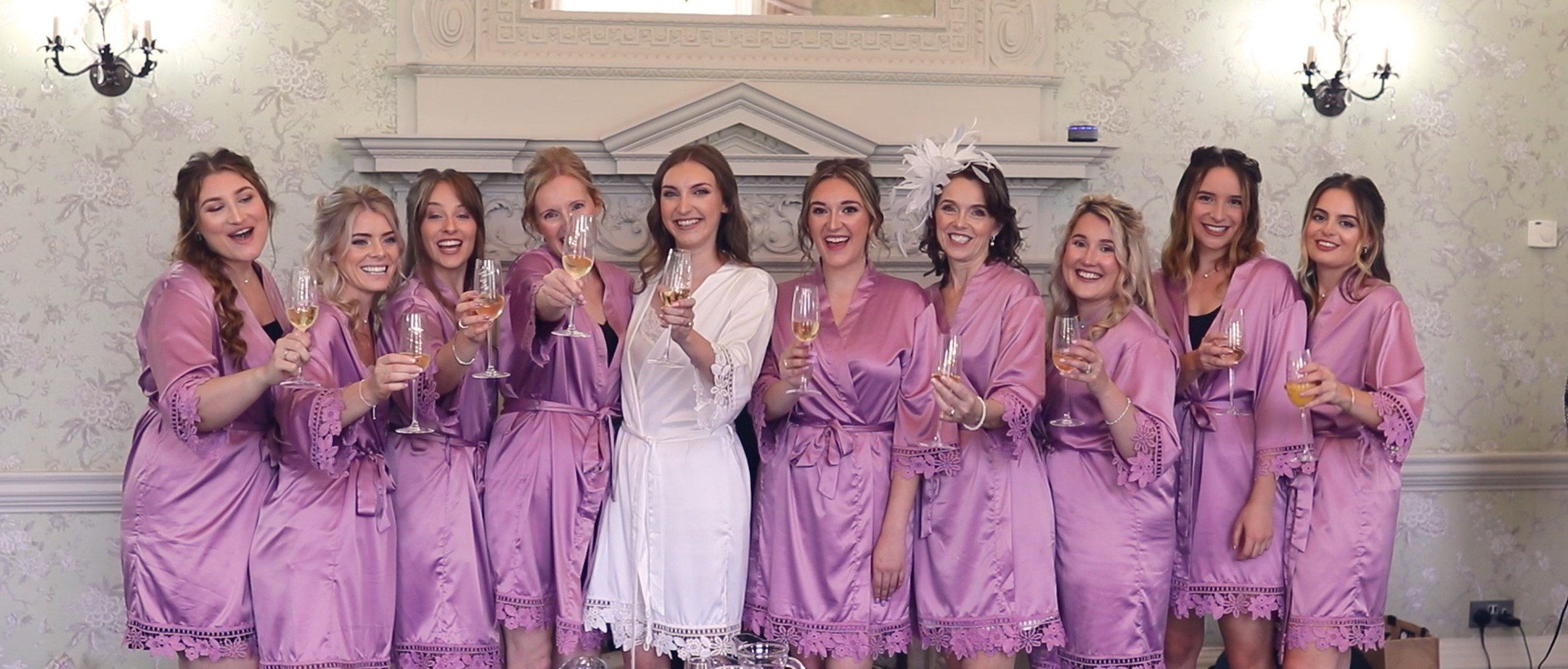 A group of eleven women in pink robes and one woman in a white robe celebrating together, holding glasses of wine or champagne, in a decorated room with floral wallpaper and chandelier lighting.