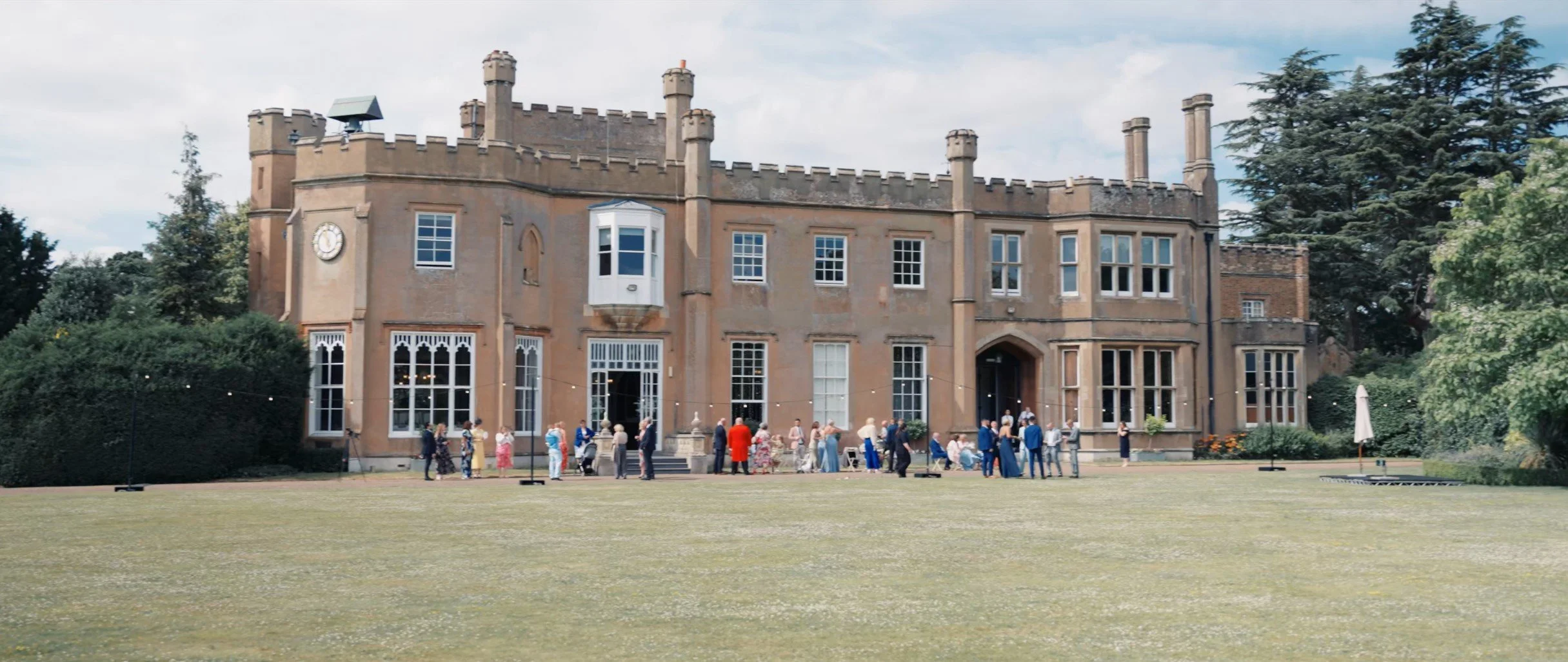 A large historic castle-like building with tan stone walls and multiple chimneys. People dressed in formal attire are gathered outside in front of the entrance, suggesting a wedding or special event. The building has a clock on the front and an outdo