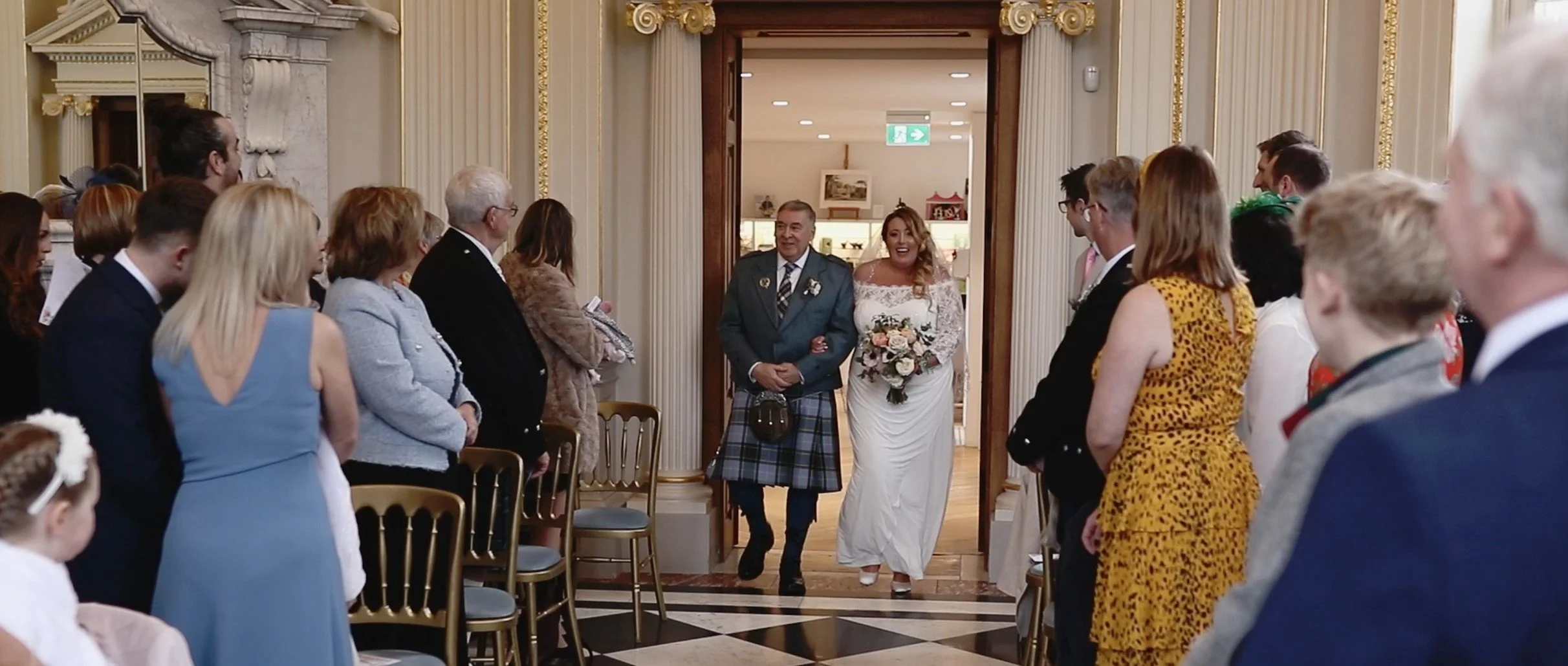 Bride walking down the aisle accompanied by a man in traditional Scottish kilt, as guests seated on both sides watch and smile inside an ornate wedding venue.