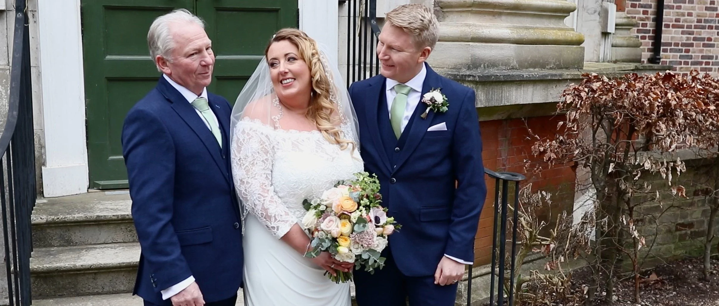 A bride in a white lace wedding dress holding a bouquet of flowers stands between two men in dark blue suits, one older and one younger, outside a building with brick and stone steps. They are all smiling and looking at each other.