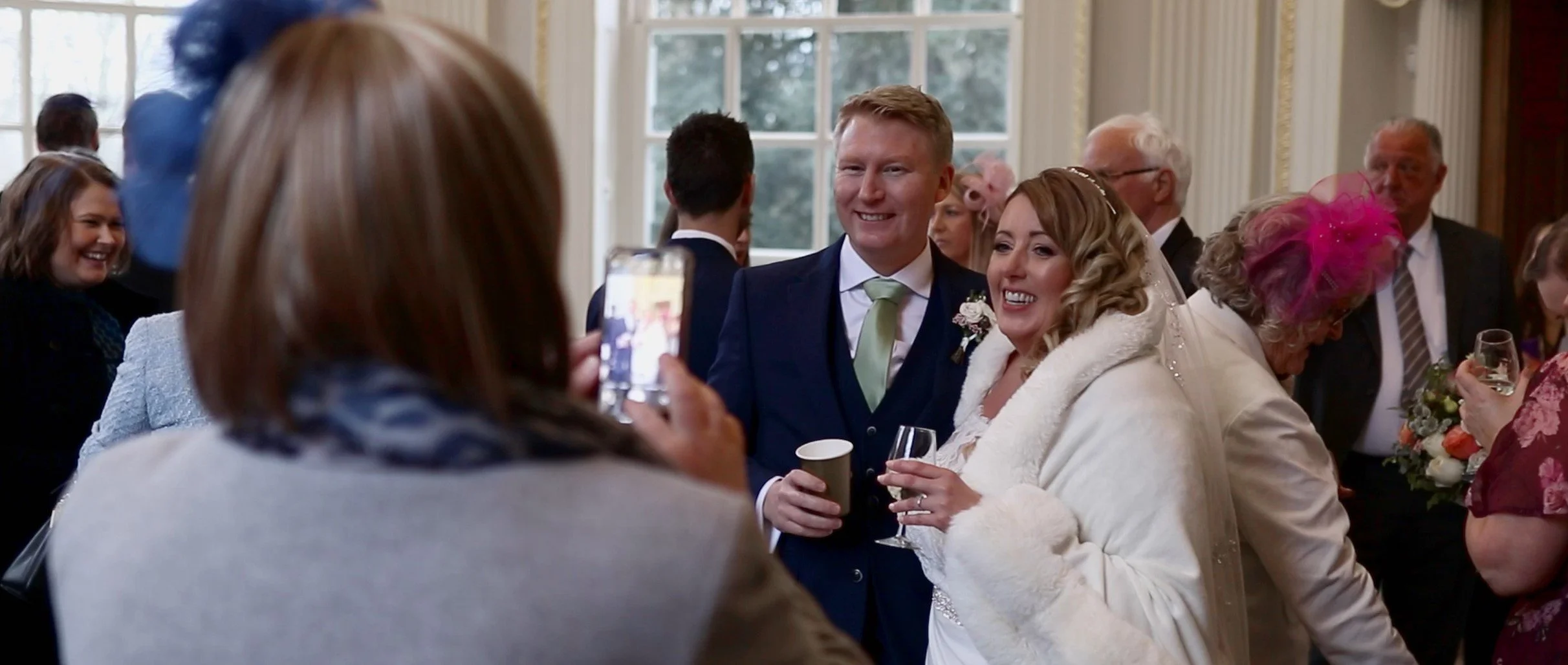 Couple smiling and taking a photo at a wedding reception with guests in the background