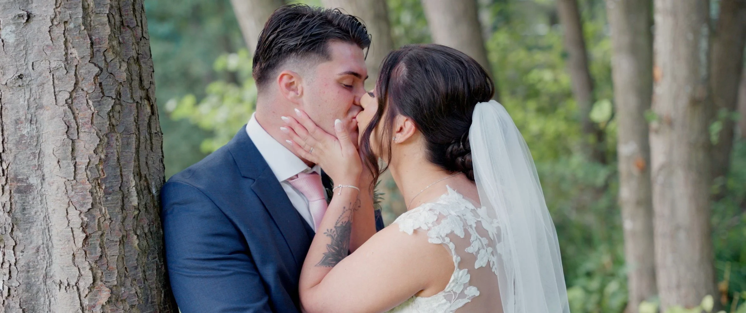 A bride and groom kiss in a wooded outdoor setting, with trees in the background. The bride wears a white lace wedding dress and veil, and has dark hair styled in an updo. The groom wears a navy suit with a white shirt and pink tie, and has short dar