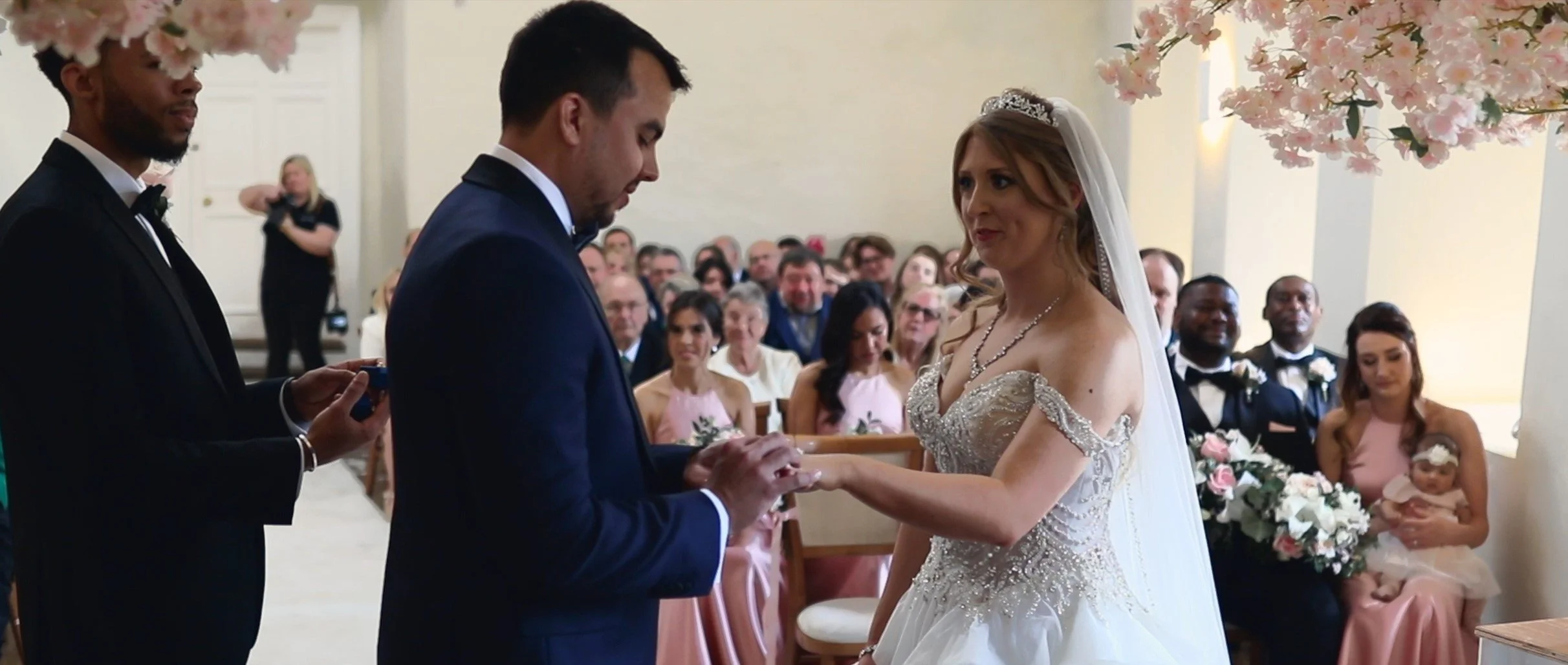 A wedding ceremony with a bride and groom exchanging rings, officiated by a man in a tuxedo, with guests seated in the background.