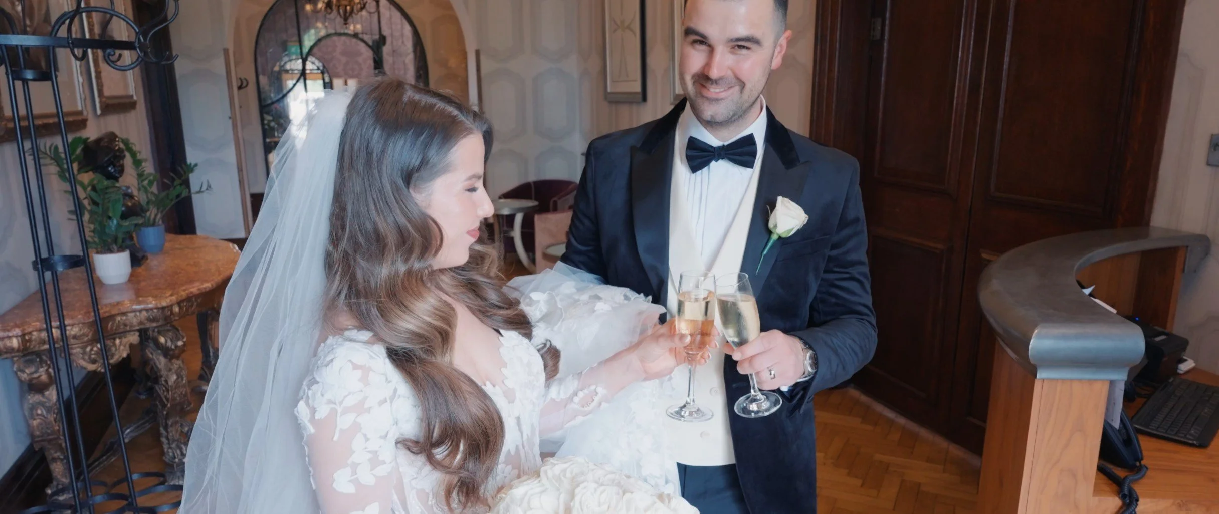 A bride and groom toast with champagne glasses inside a decorated room, smiling and celebrating their wedding.