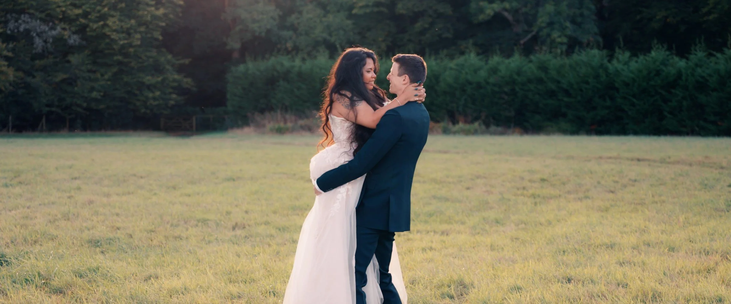 A couple in wedding attire embracing in a grassy field during sunset, with trees in the background.