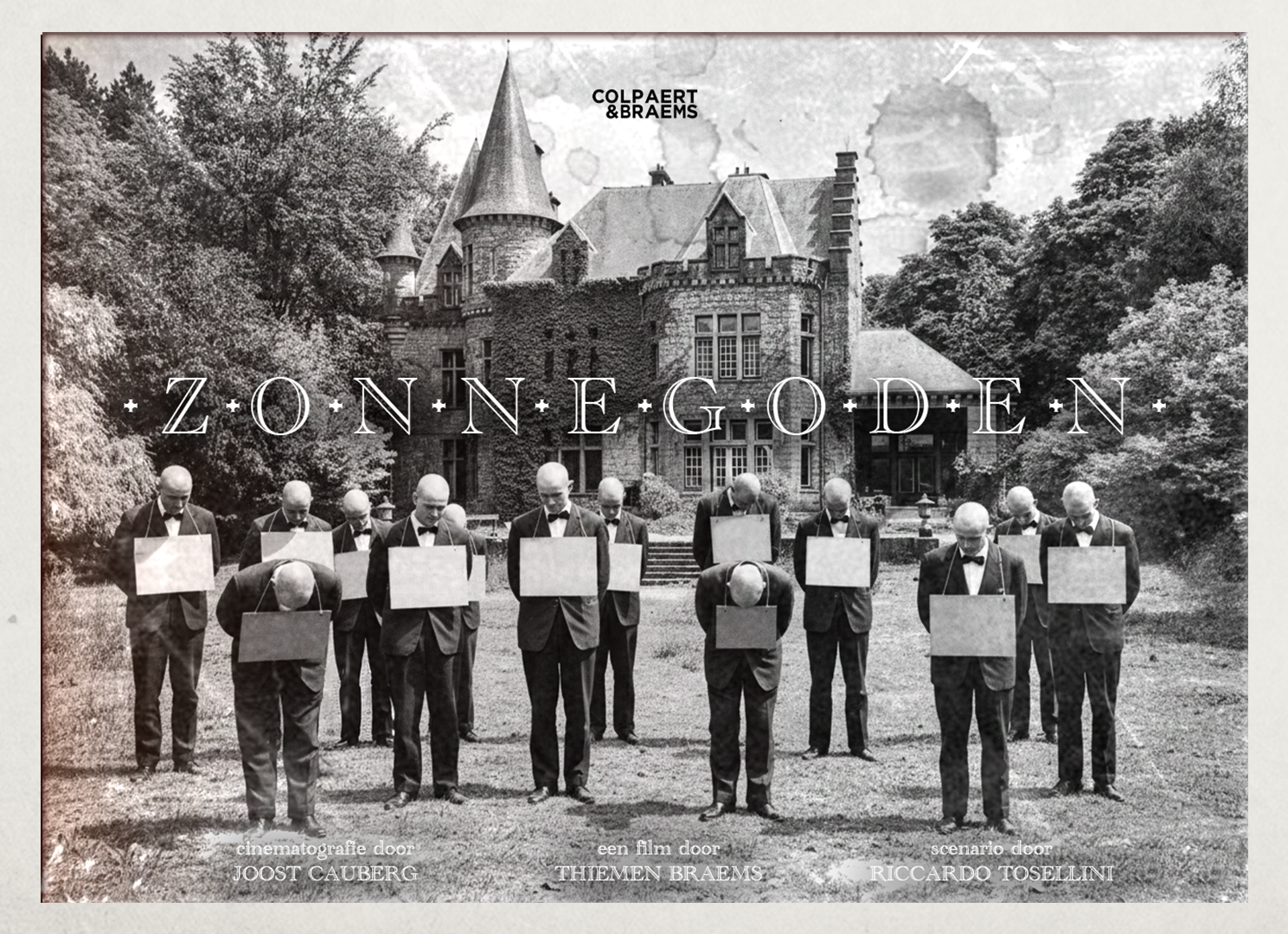 Zwart-wit foto van een groep mannen in formele kleding, met hun hoofden naar beneden, staand voor een oud kasteel, met het woord 'ZONNEGODEN' over de afbeelding.