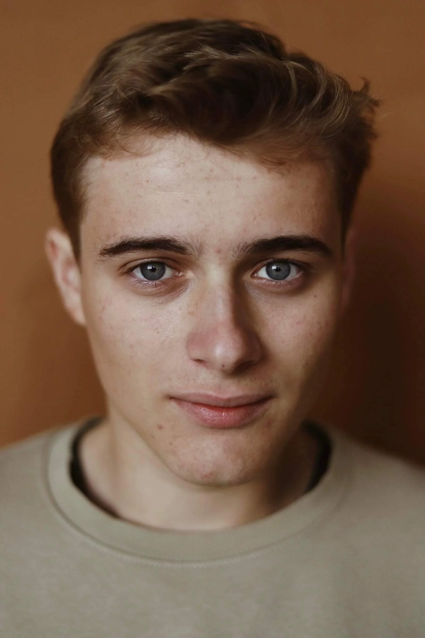 Close-up van een jonge man met blauwe ogen en roodbruin haar, in een beige trui, tegen een bruin achtergrond.