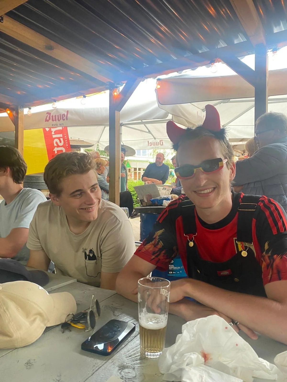 Mensen zitten aan een tafel onder een overkapping in een buitenruimte, met één persoon die draagt ​​​​hond-oren en zonnebril, terwijl anderen lachen en praten.