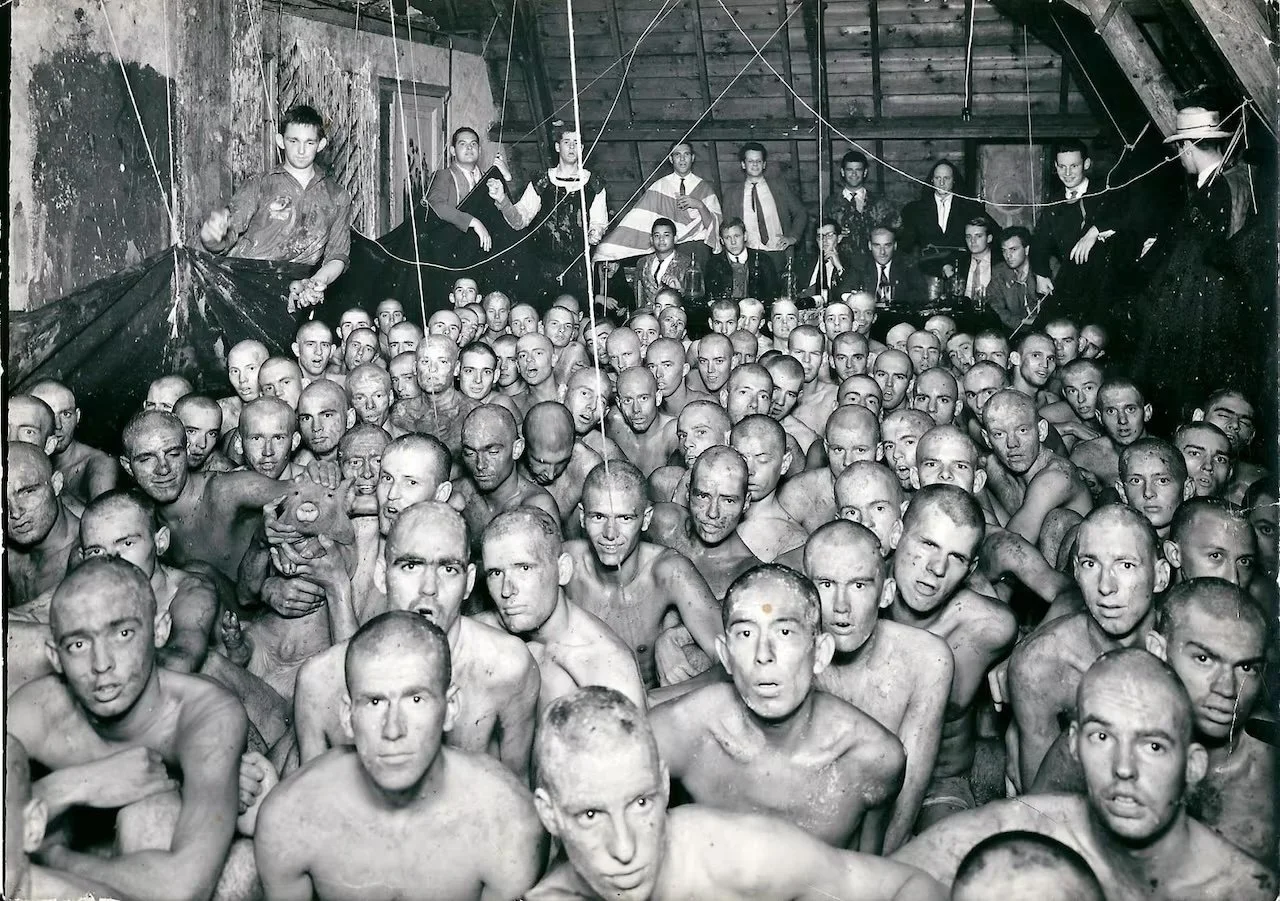 Zwarte en witte historische foto van een grote groep mensen zonder kleren, voornamelijk zeer jonge mannen, verzameld in een houten zaal met mannen op een verhoogde plank met vlaggen achter hen.