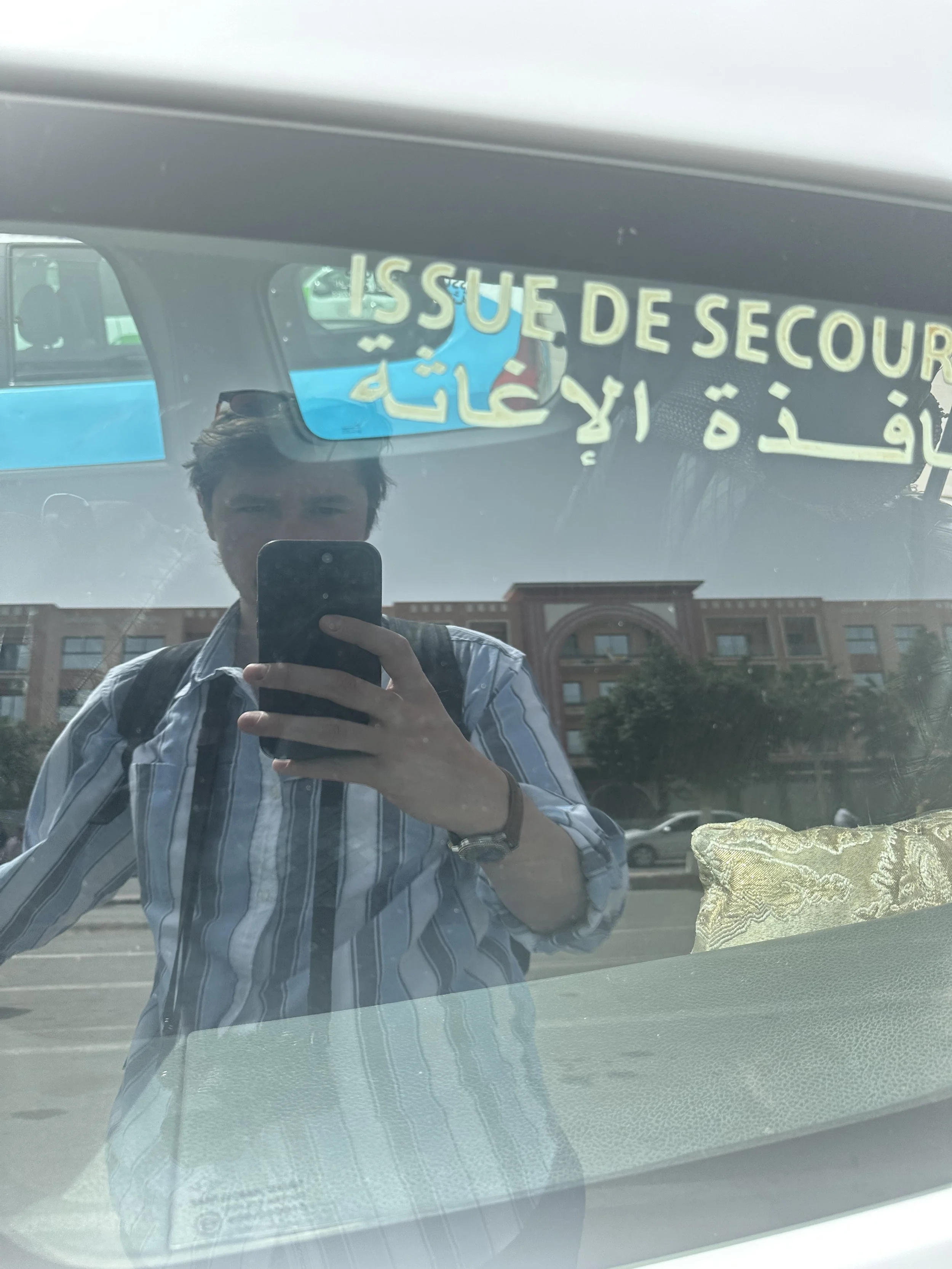 Reflexion eines Mannes mit Streifenhemd und Backpack, der ein Smartphone zur Aufnahme benutzt, in einem Fahrzeug mit evidenter Markierung "ISSUE DE SECURITÉ" auf der Windschutzscheibe, mit einem Gebäude im Hintergrund.